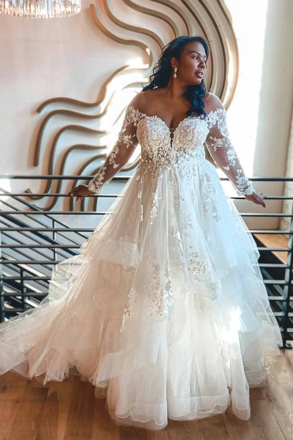 A woman in a wedding dress standing indoors by a railing, with a modern wooden wall art piece behind her and sunlight coming through a window on the right.