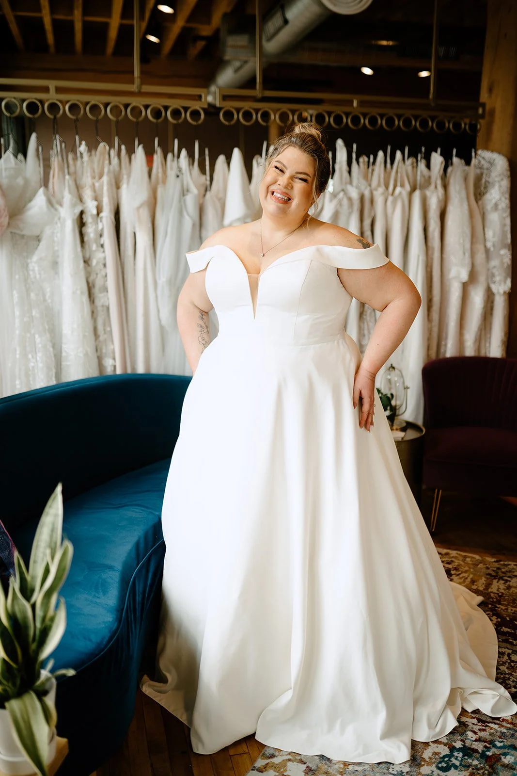 A woman in a white wedding dress smiling in a bridal boutique with a backdrop of wedding gowns.