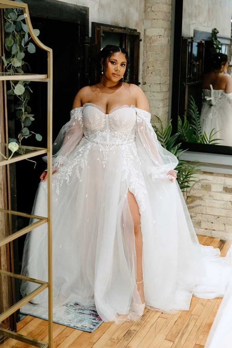 A woman in a white wedding dress with off-the-shoulder sleeves and a slit in the skirt stands inside a room with a brick wall, a mirror, and green plants. She is posing and looking at the camera.