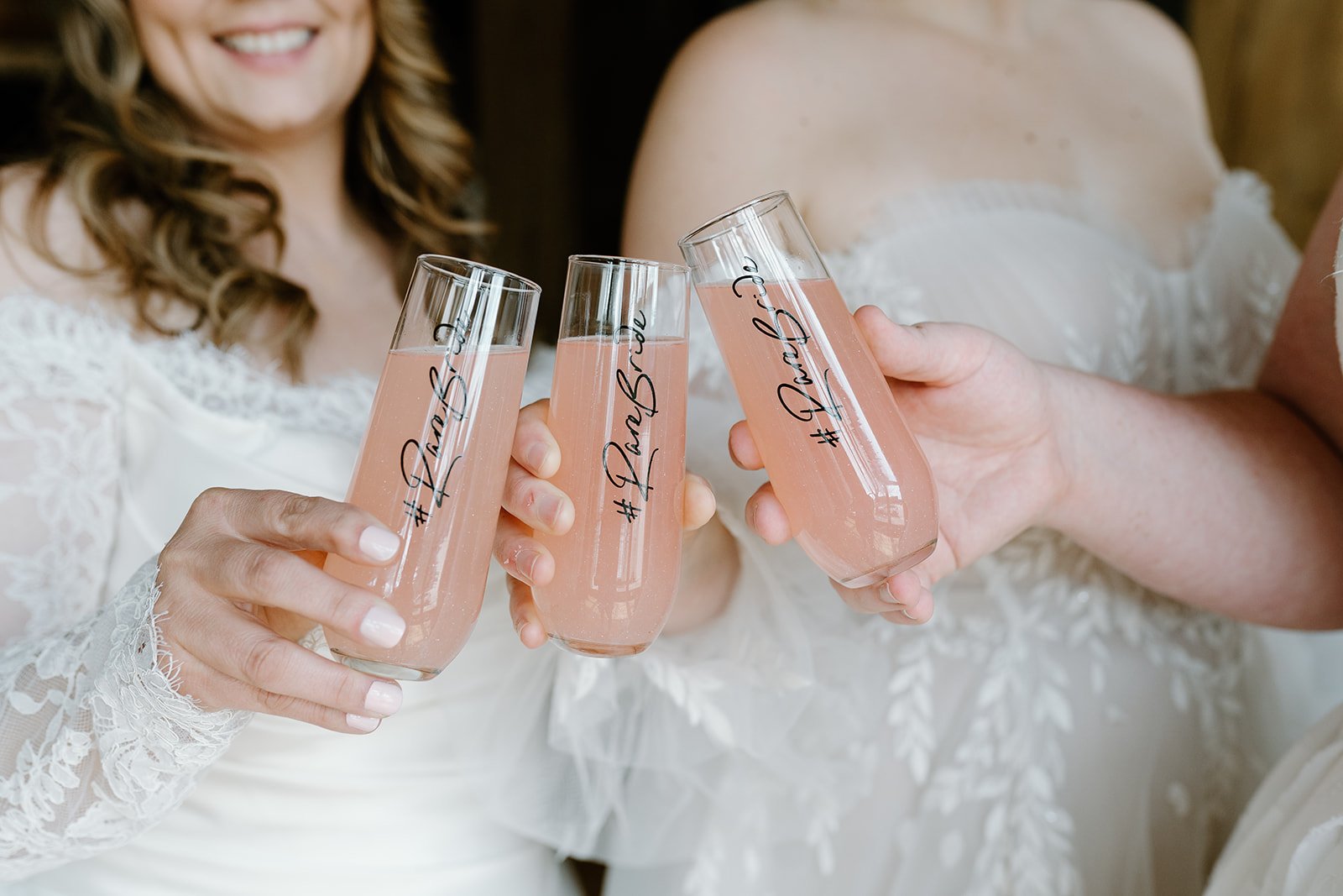 Two women in wedding dresses clinking glasses of pink champagne with hashtag "RareBride" written on the glasses.