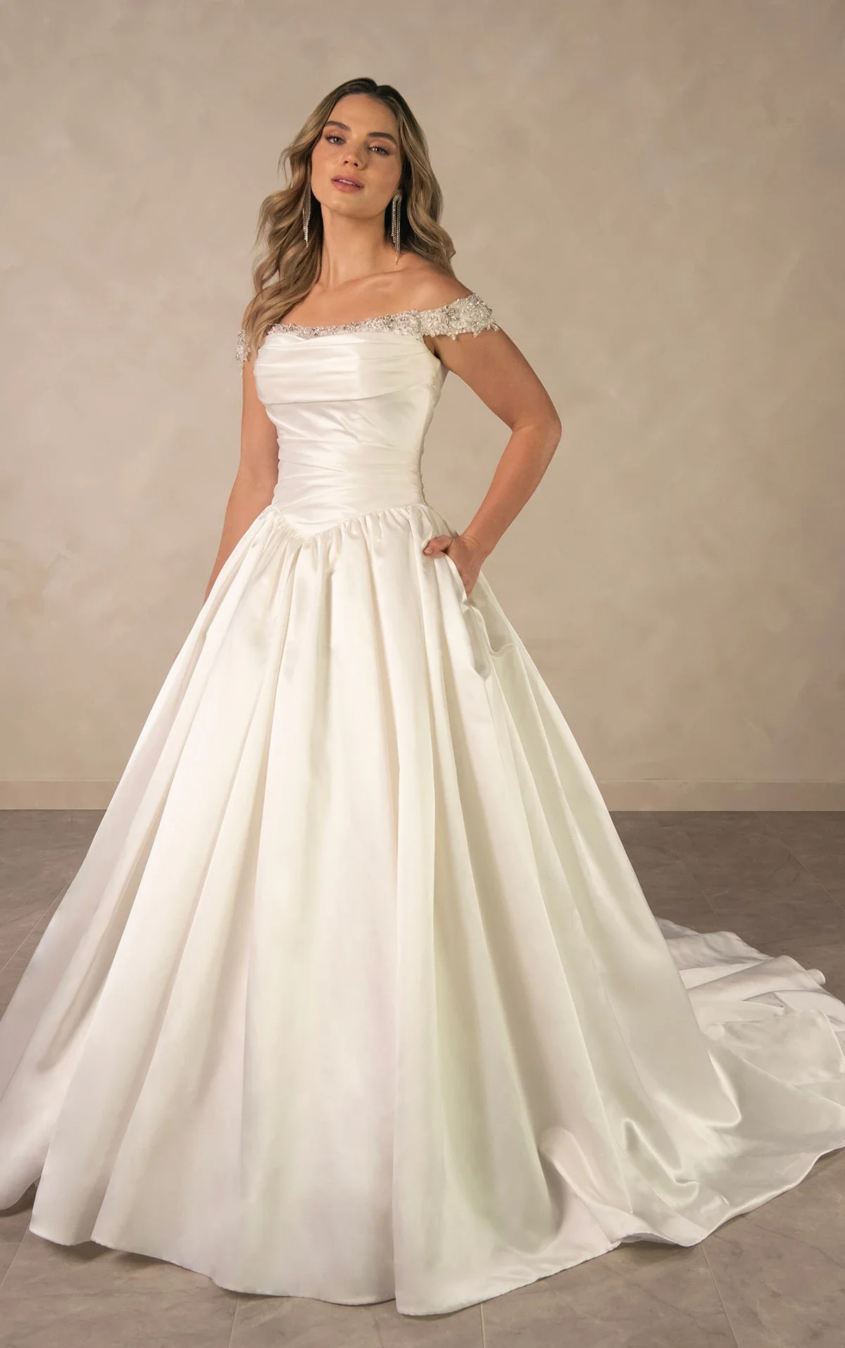 A woman in a white satin off-shoulder wedding gown with beaded embellishments on the neckline and waist, posing with one hand on her hip and the other hanging down, against a plain beige background.