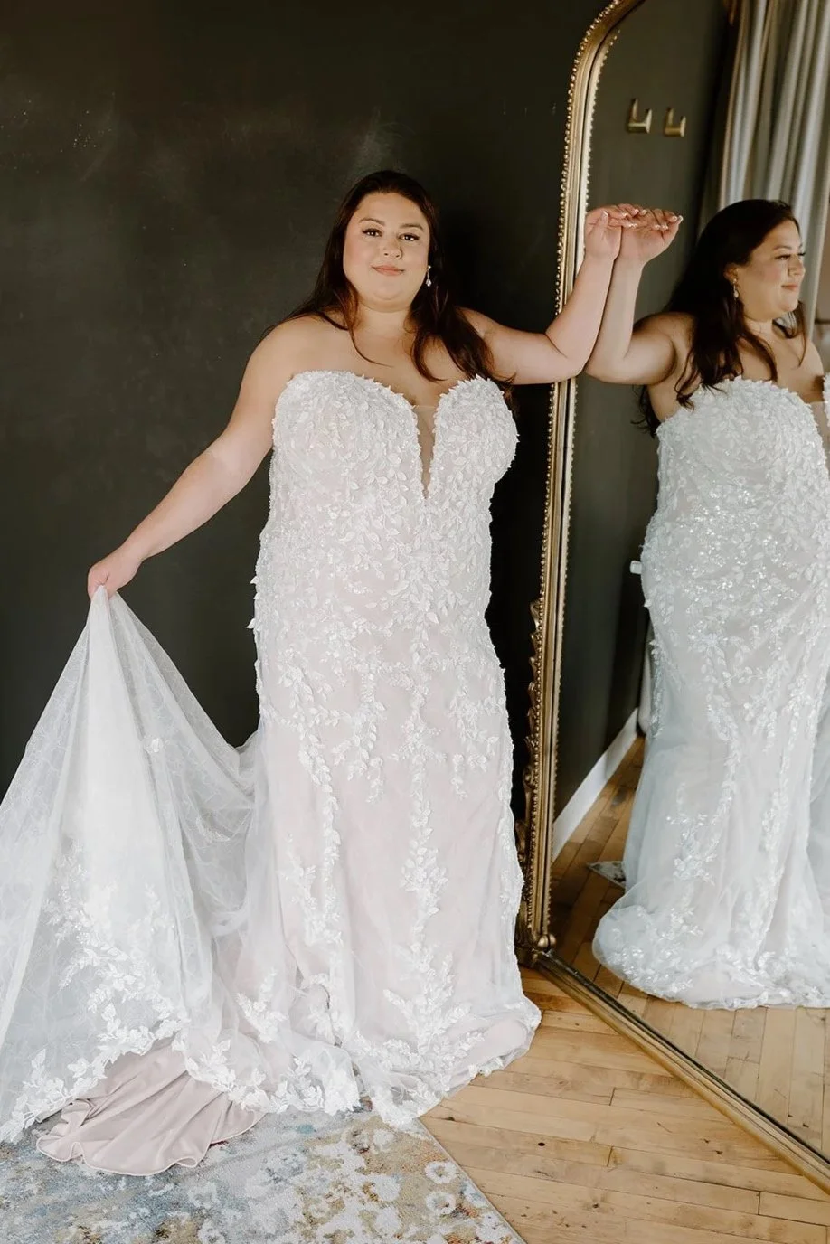 A woman in a white wedding dress with floral embroidery, holding the skirt out with her left hand, standing in front of a mirror.
