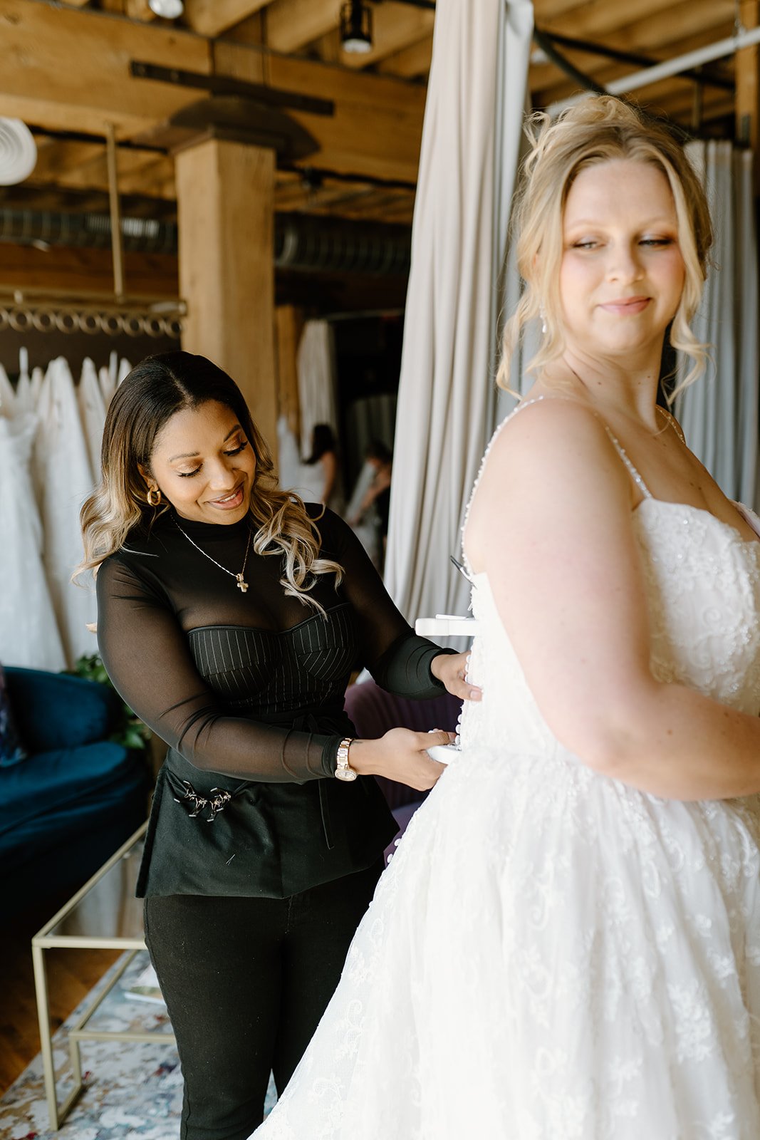 A woman in a white wedding dress is smiling as another woman helps her lace up the back of her gown. The second woman is wearing a black top and black pants and is smiling while assisting.