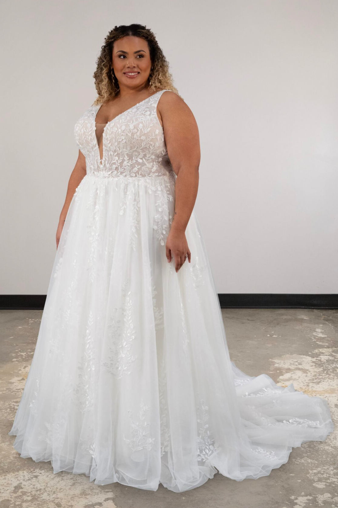 Woman in a white wedding dress with lace and floral details, standing indoors in front of a plain light-colored wall.
