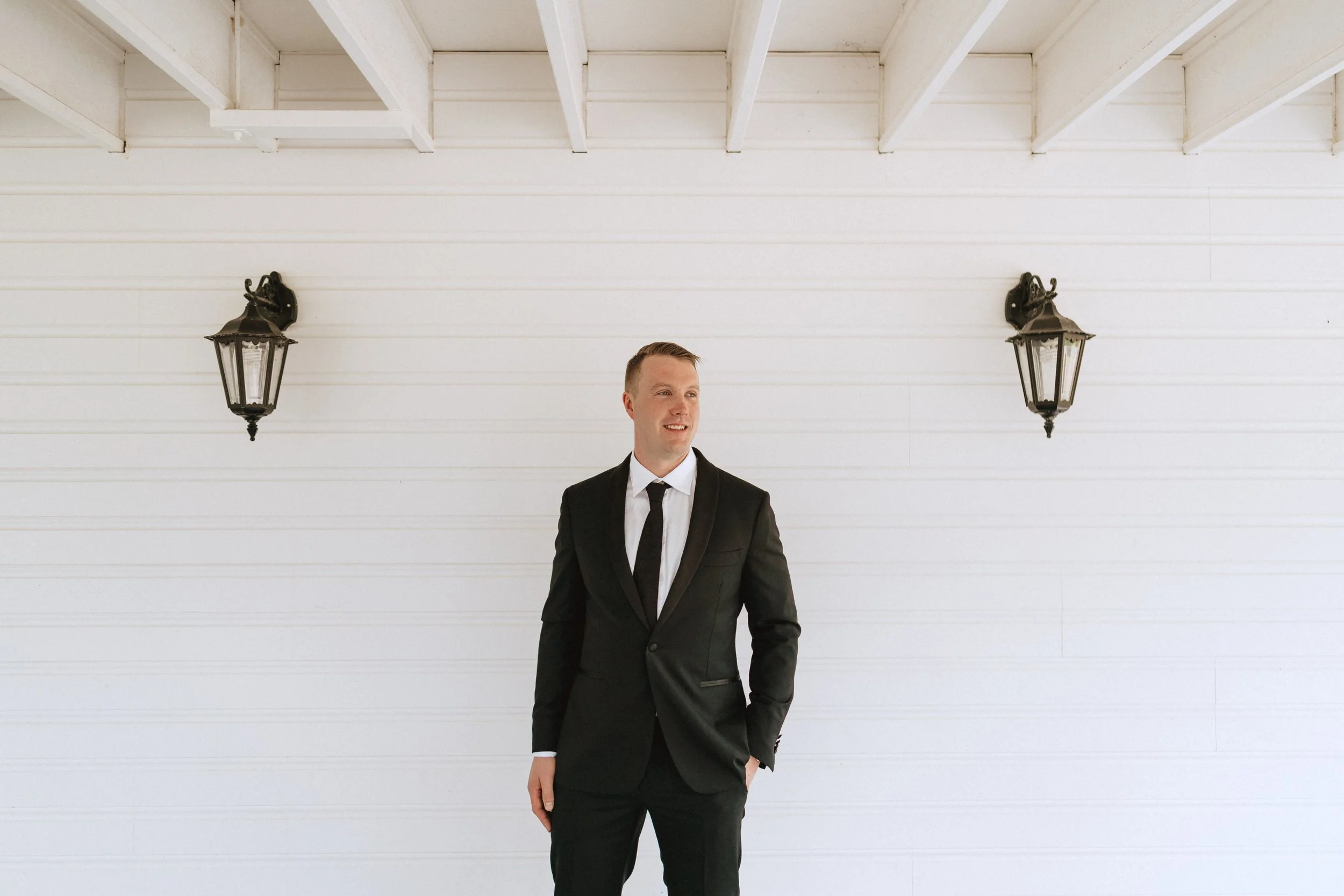 Groom standing up against a wall in his suit looking out to ceremony.