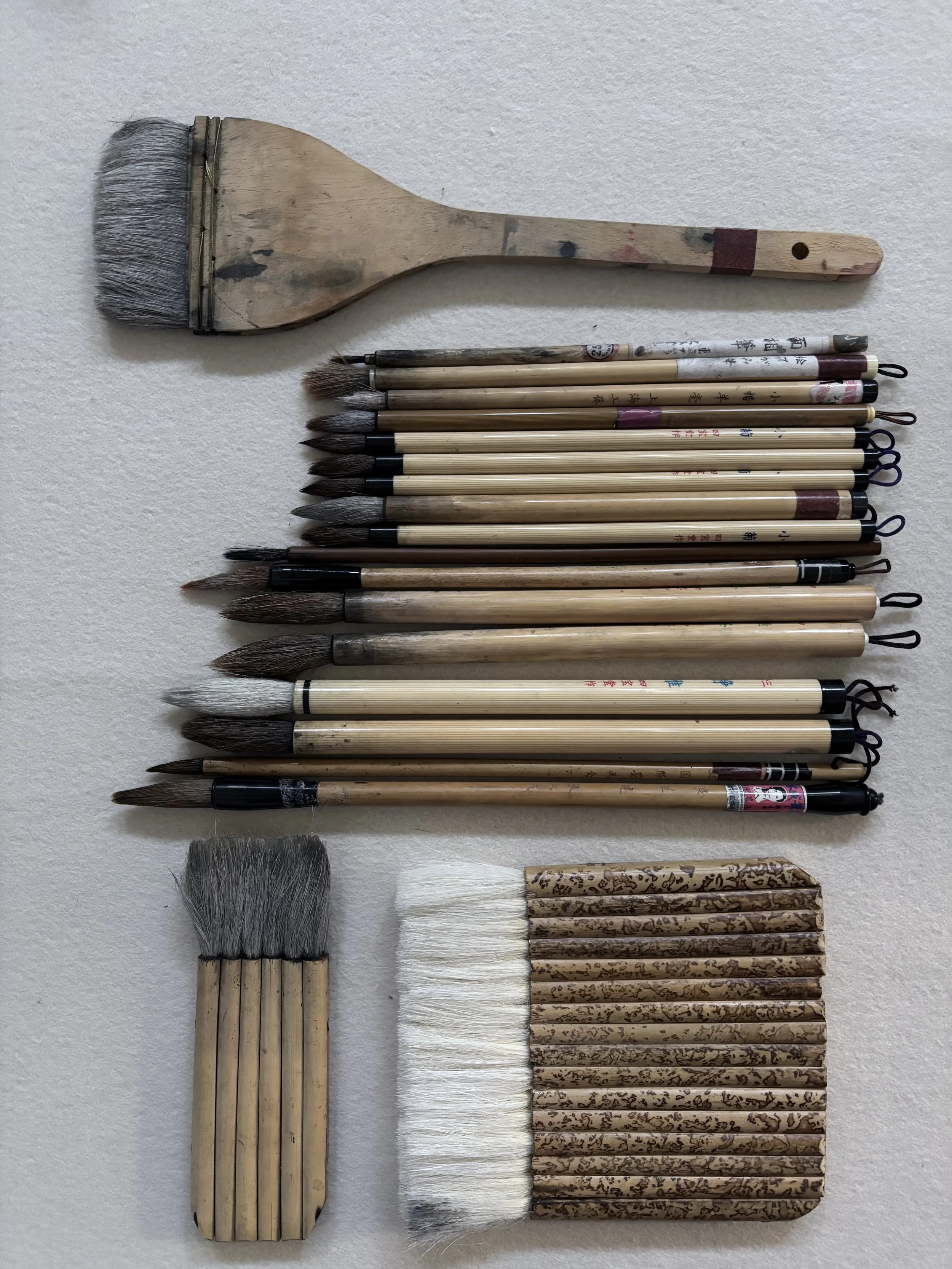 Collection of traditional Asian calligraphy brushes and a large flat brush for painting, arranged on a white surface.