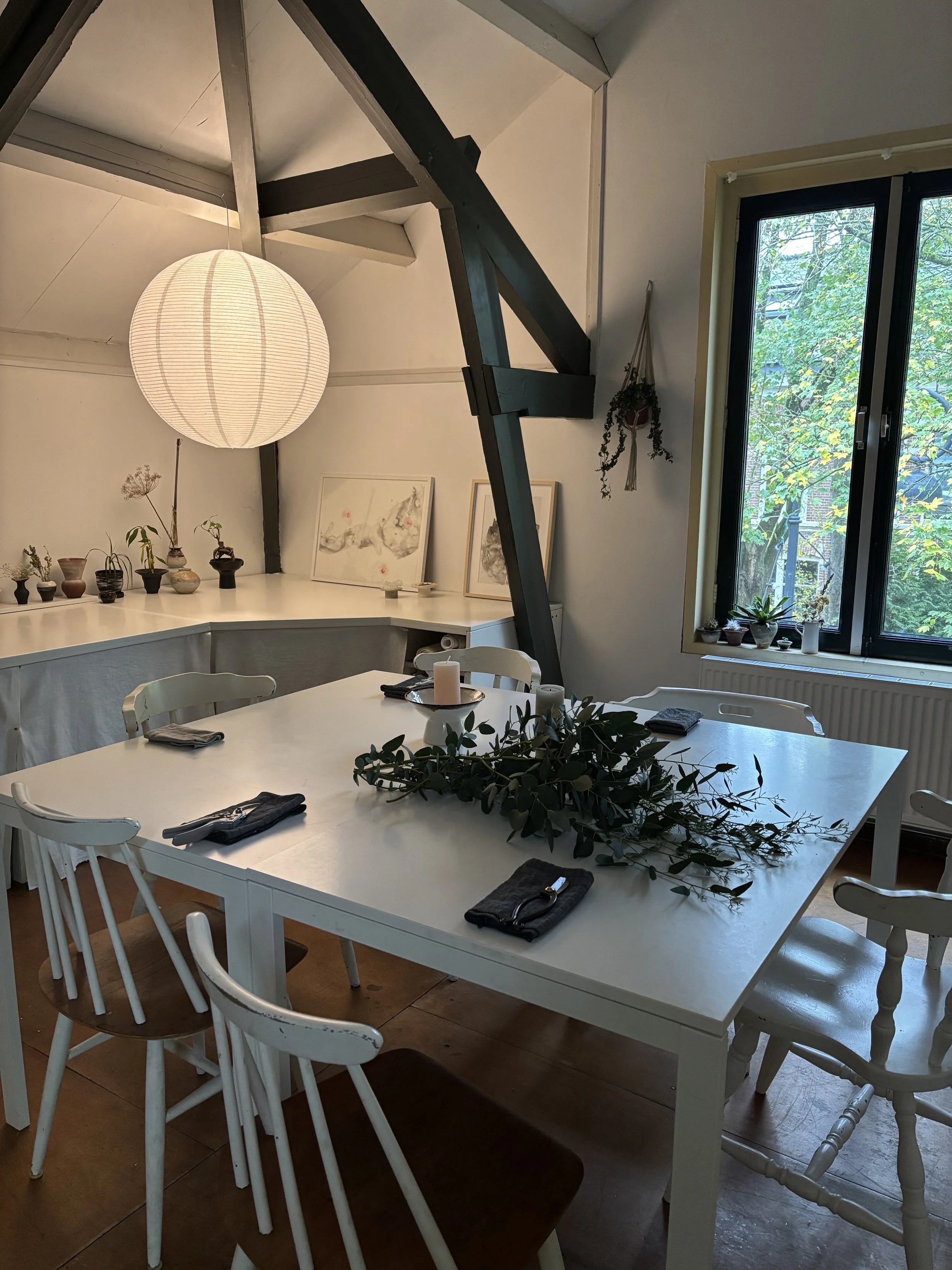 A dining room with a white table set with napkins and utensils, surrounded by mixed chairs, a centerpiece with greenery, a large window with a forest view, potted plants on the window sill, and artwork on the back wall, with a hanging paper lantern and decorative wall hanging.