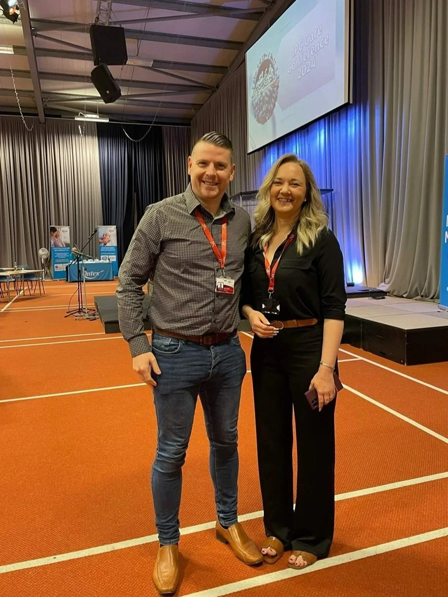 Neil Fossett (EMT) & Rachel Holden (Paramedic) Two smiling conference attendees standing in front of a stage and a large screen showing the text "IPC Care Conference 2024" at an indoor event.