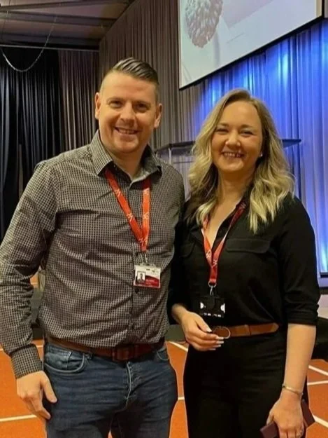 Neil Fossett (EMT) & Rachel Holden (Paramedic) Two smiling conference attendees standing in front of a stage and a large screen showing the text "IPC Care Conference 2024" at an indoor event.