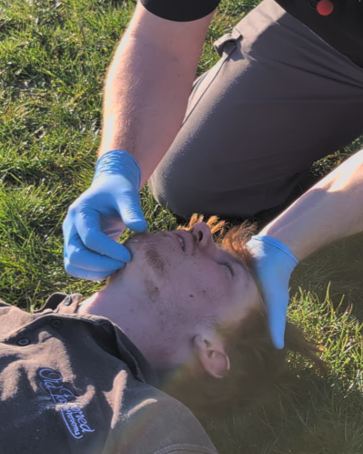 Person performing CPR or first aid on an injured individual lying on the ground in a grassy area, wearing gloves.