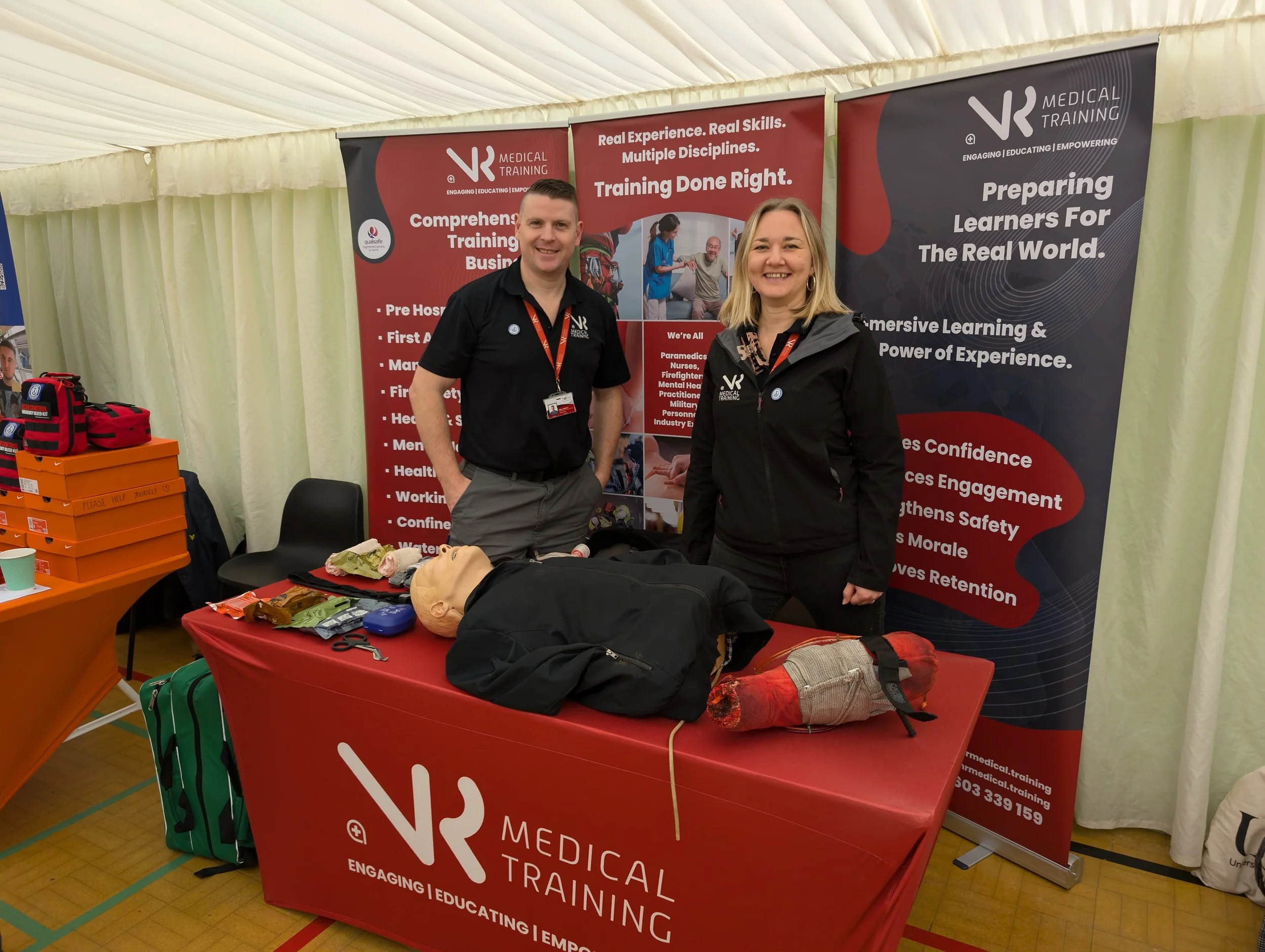 Rachel Holden & Neil Fossett standing behind a display table at a medical training booth, with de-activated medical mannequins and training supplies, in a large enclosed event space with banners promoting medical training programs.