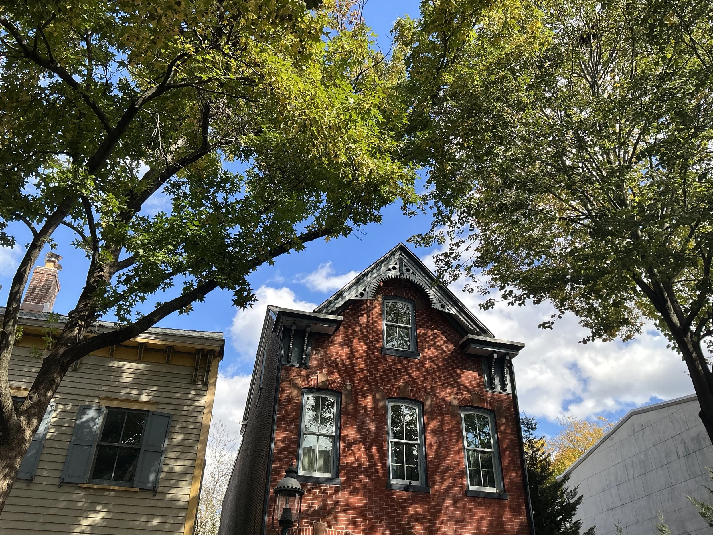 A red brick house with black window frames, partly obscured by green trees, under a blue sky with some clouds.
