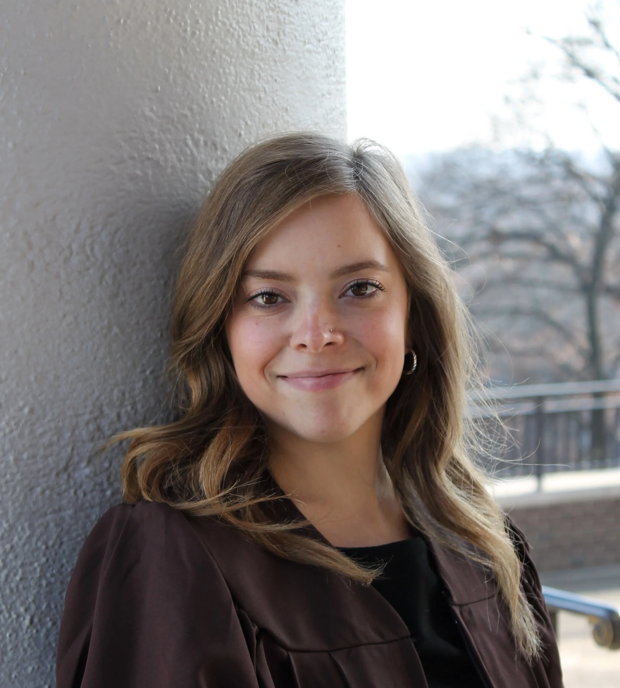 A young woman with shoulder-length wavy hair smiling and leaning against a textured wall outdoors during daytime.