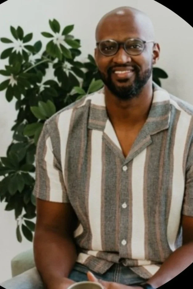 A man with glasses, a beard, and a shaved head smiling while sitting in front of a green leafy plant, wearing a striped short-sleeve button-up shirt.