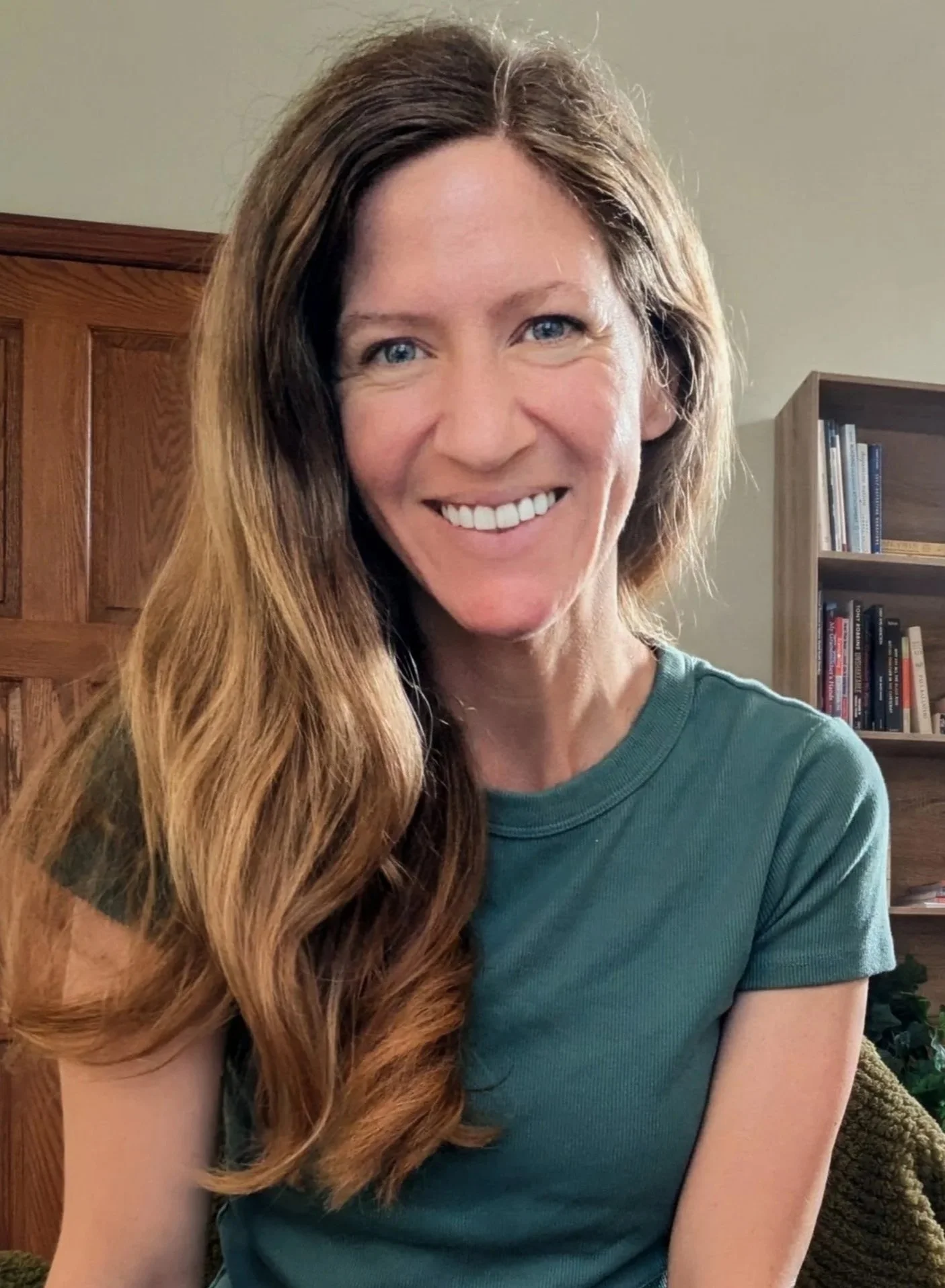 A woman with long, wavy red hair smiling at the camera, sitting on a green couch near a window.