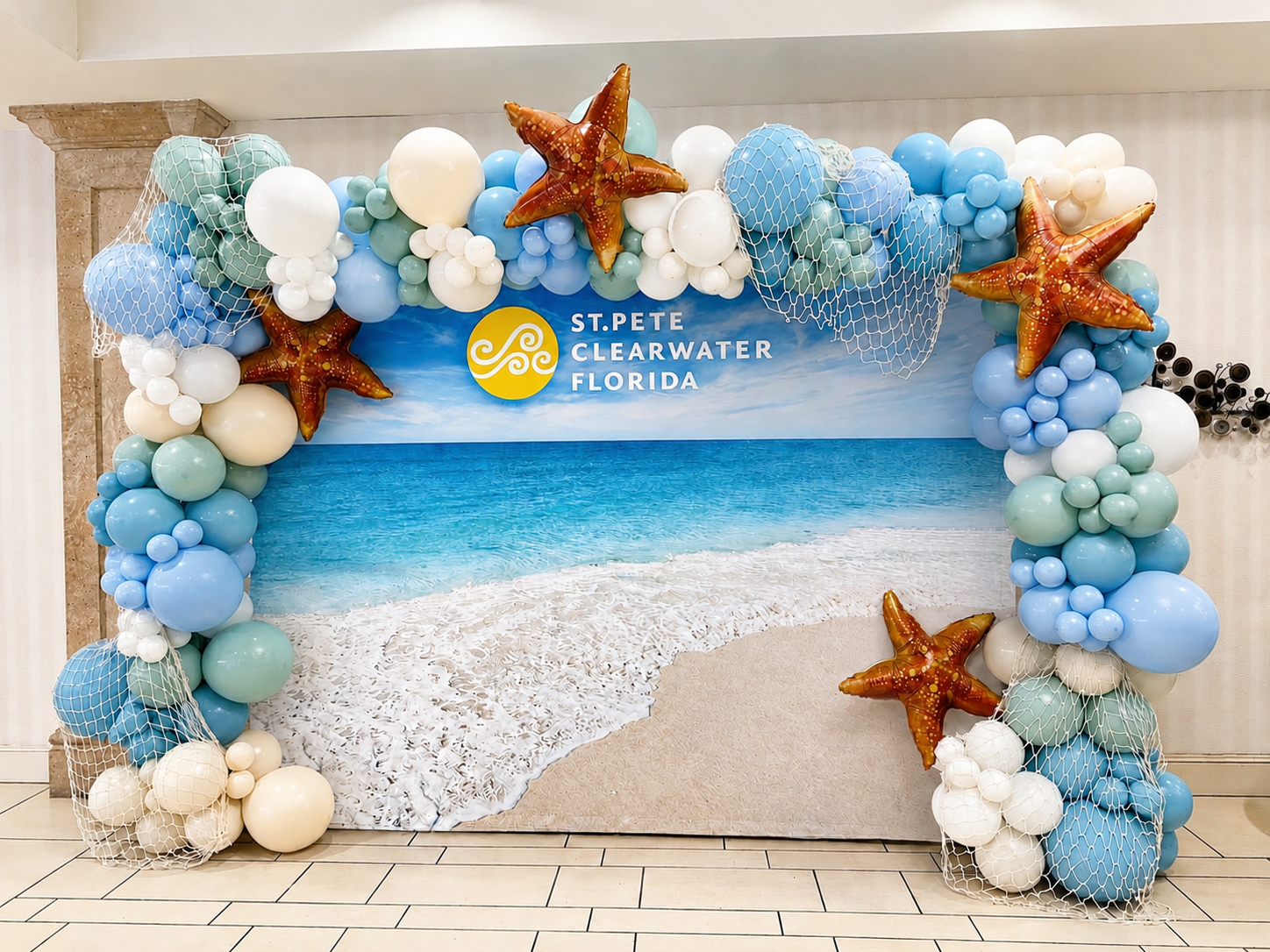 Beach-themed balloon backdrop featuring an organic garland in shades of blue, aqua, white, and sand, accented with decorative fishing nets and oversized starfish balloons.