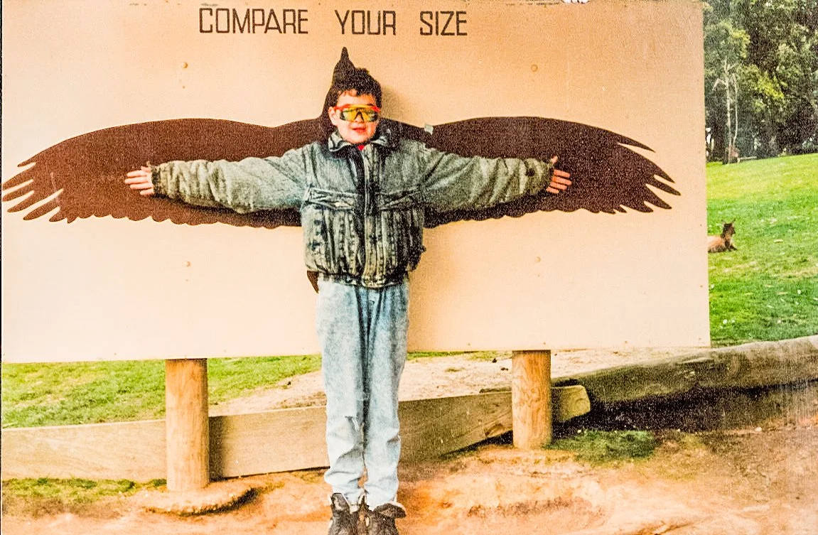 Child standing in front of a "Compare Your Size" sign with extended wings, arms spread wide, wearing sunglasses and a jacket, outdoor background with grass and trees.