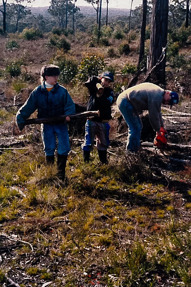 Three people, including children, collecting firewood in a forested area. One adult is using a chainsaw.