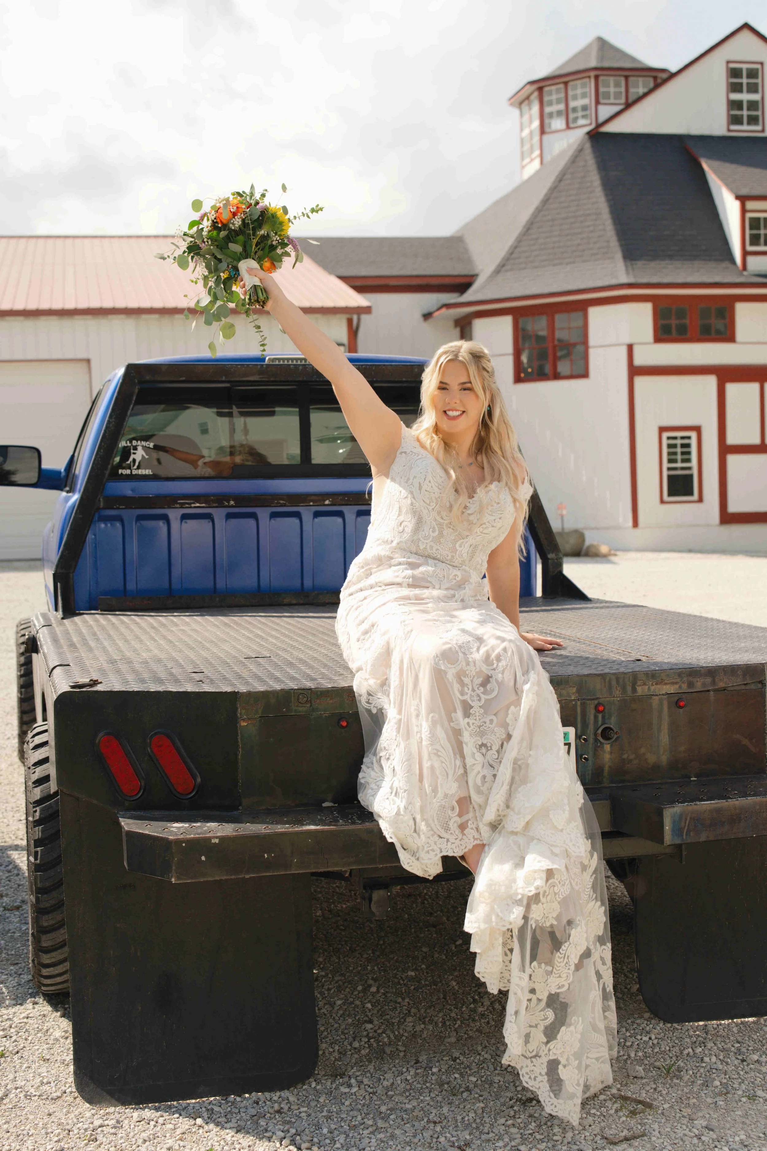 Bride posing with Grooms truck.  She is sitting on the back of the flatbed raising up her bouquet in celebration with the Governor's Pavilion in the background
