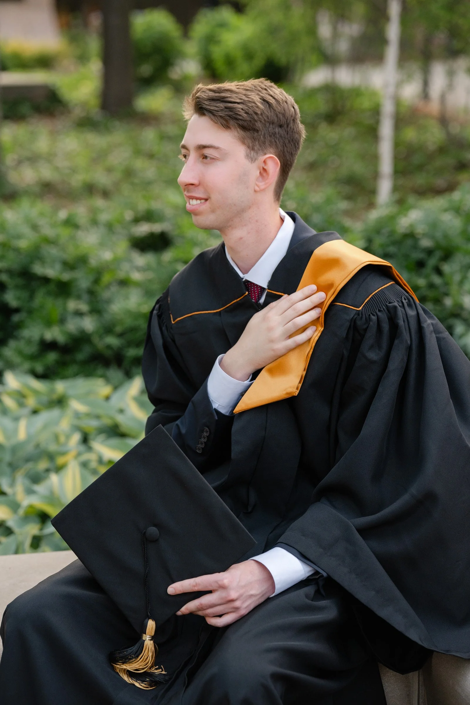 Man sitting on bench wearing Purdue Graduation gown holding his cap and stole looking off into the distance.  Photo by Heather Corbin Photography