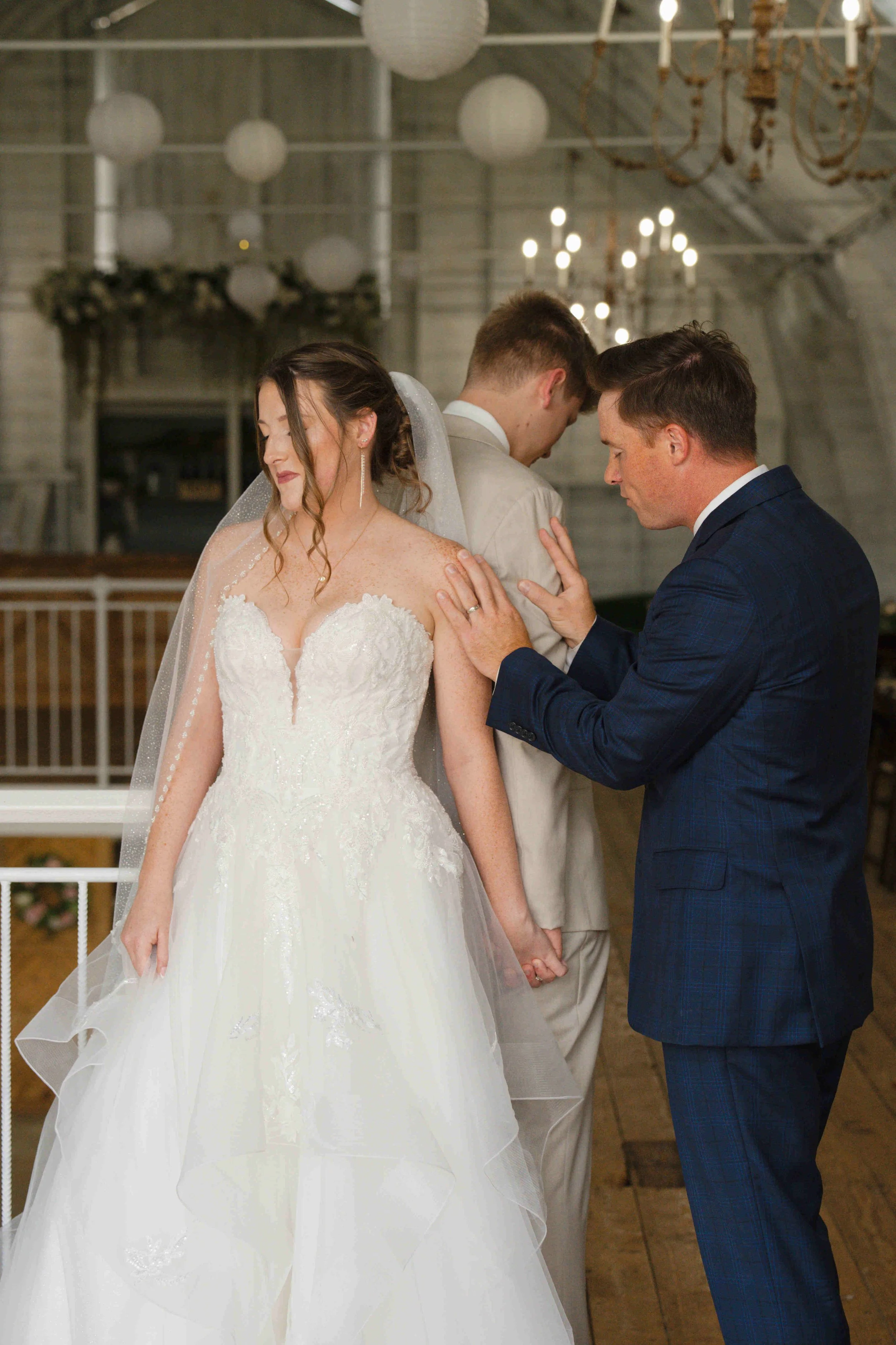 The bride and groom standing back to back holding hands for a "first touch" while their pastor prays a blessing over them.  Wedding at The Blossom Barn, Mulberry Indiana.