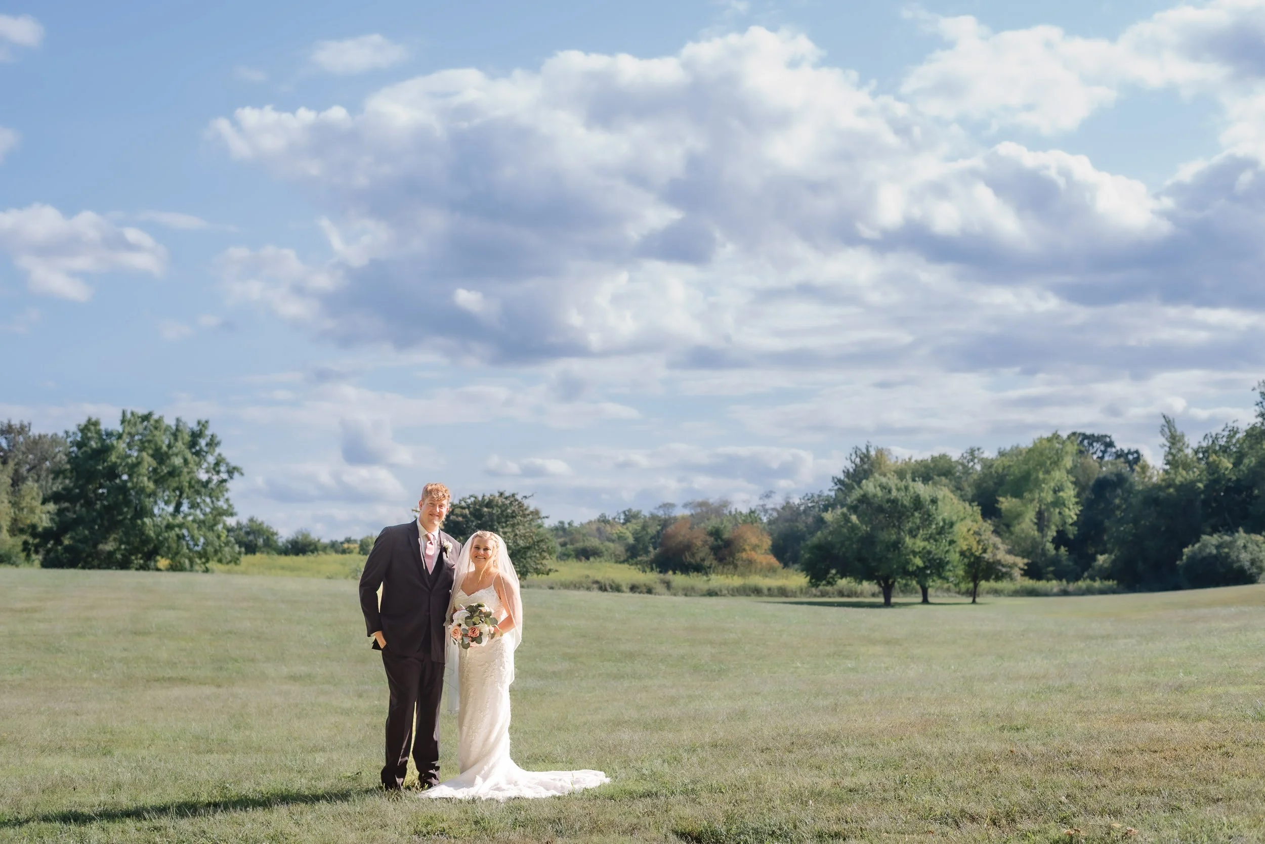 Bride and groom standing in a wide open green field with a big sky filled with puffy clouds at The Stables Event Center, Lafayette Indiana.  by heather corbin photography