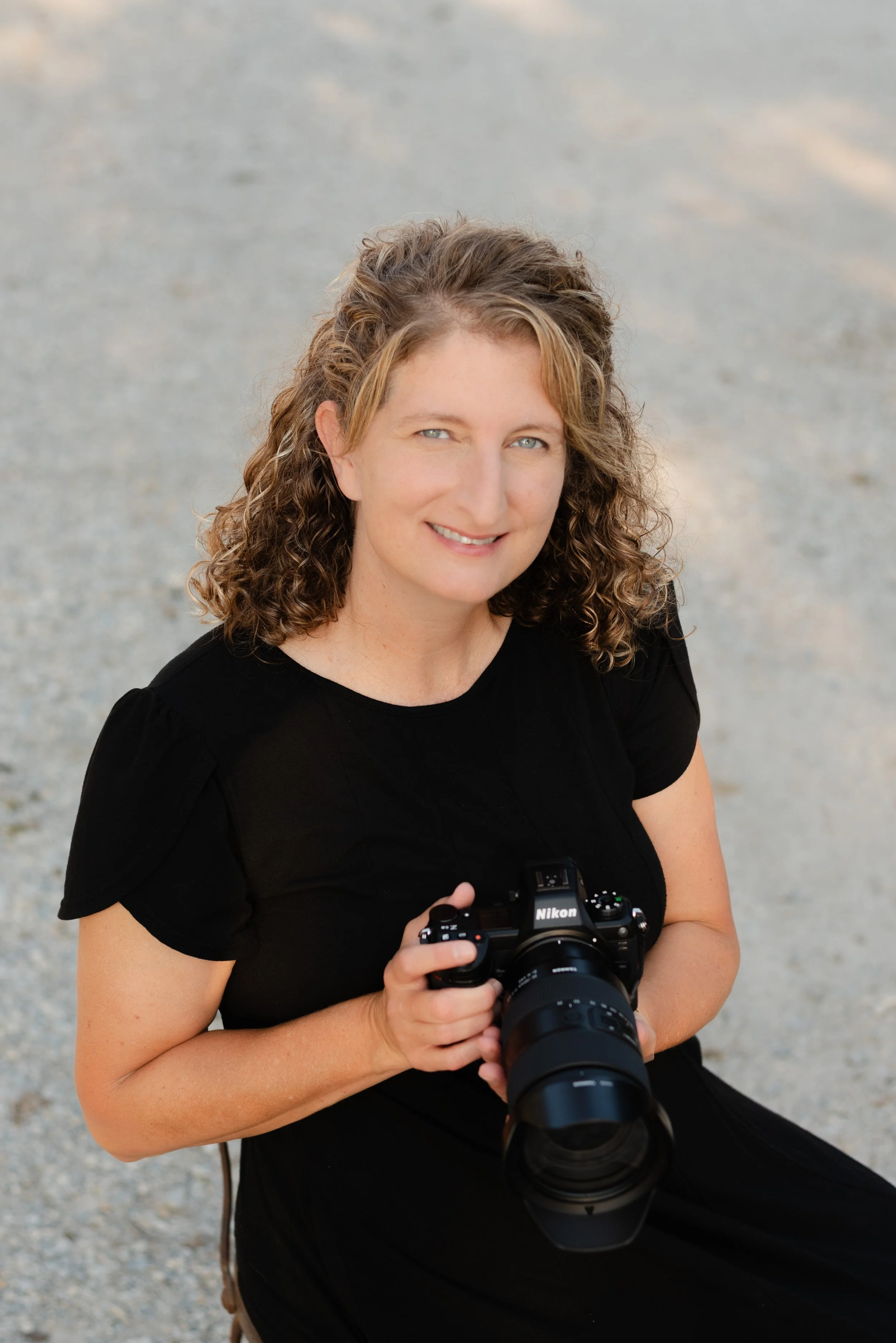 Headshot of Heather Corbin of Heather Corbin photography.  Black dress neutral background.  holding nikon camera