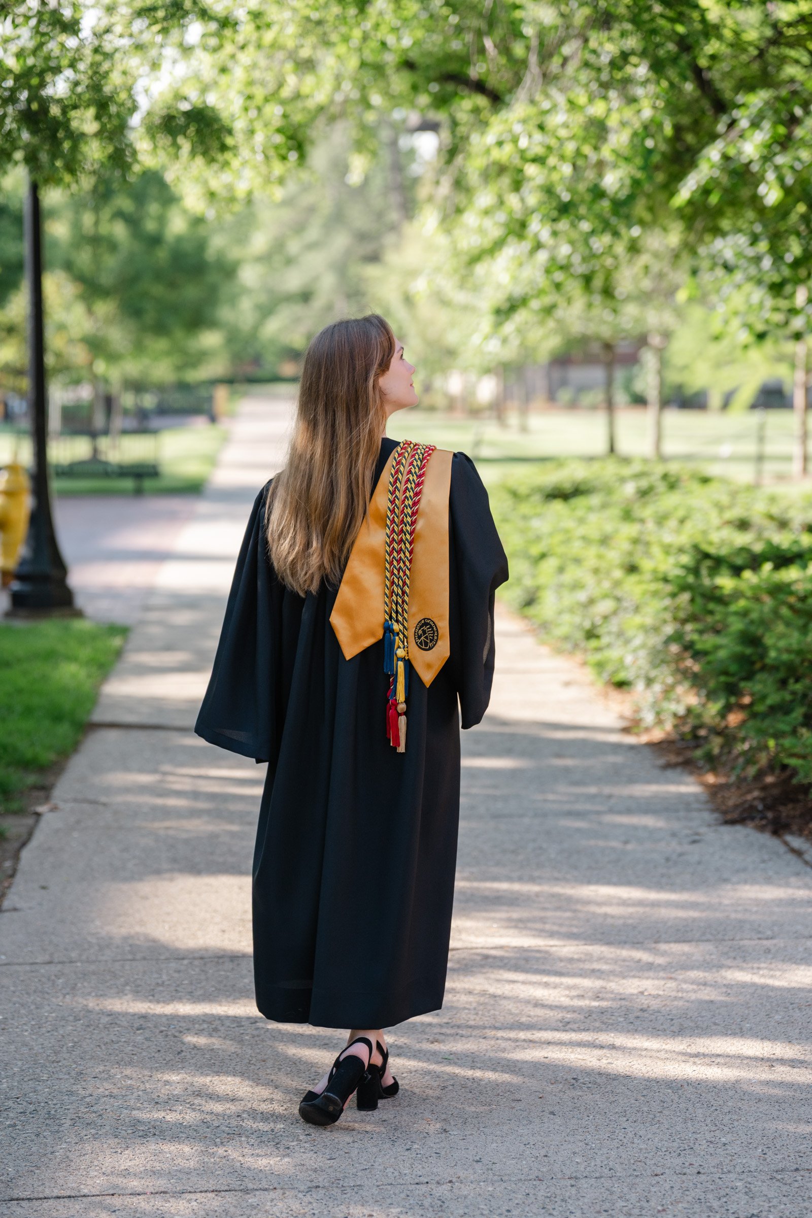 Purdue Cap and Gown session woman facing away from the camera looking off her right shoulder. wearing her graduation gown with her stole and cords over her shoulder.