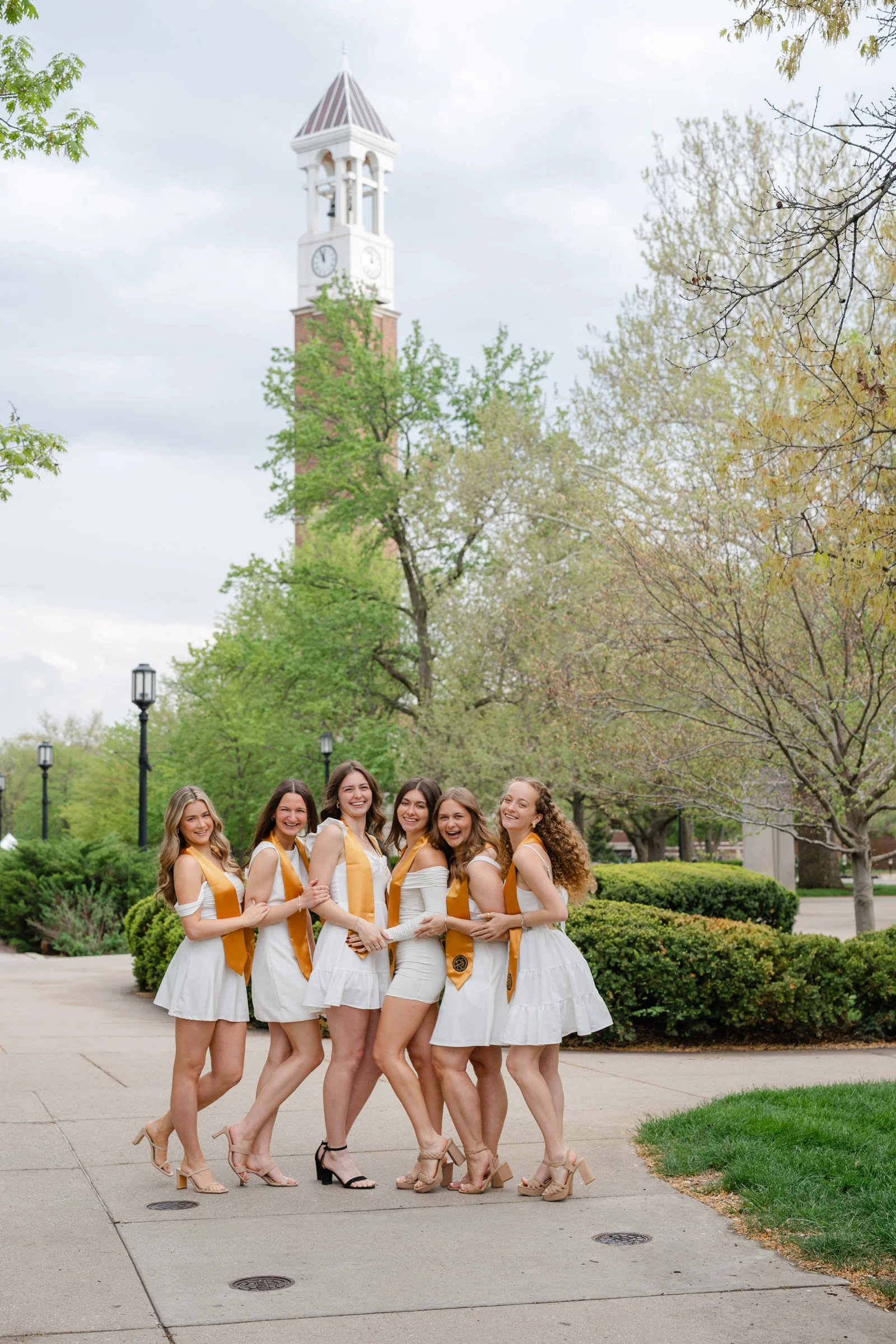 Group of Purdue nursing graduating friends posing together for a picture.  The Purdue Bell tower is in the background.  The girls are all wearing white dresses.  Photo by Heather Corbin Photography.