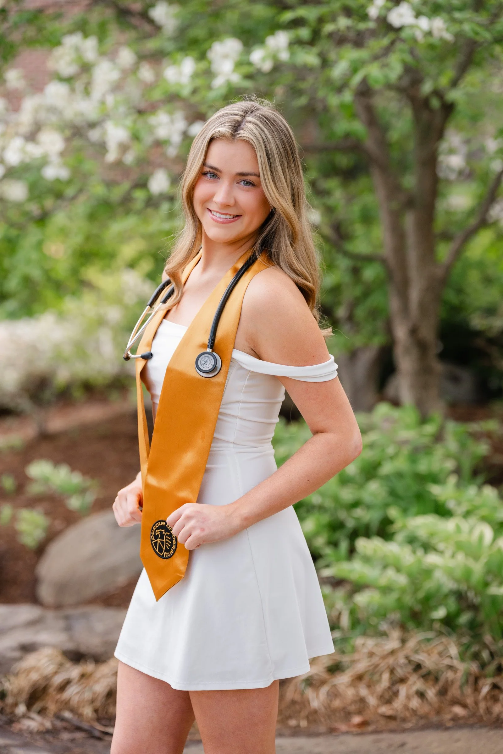 Purdue Nursing Student in white dress with Purdue gold stole and stethoscope around her neck.  Flowering trees in background.  purdue graduation photo by Heather Corbin Photography