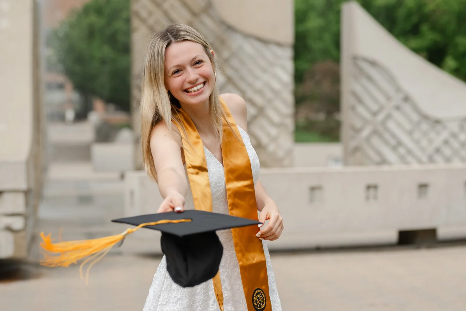A woman in white dress with Purdue Graduation Cap tossing cap during graduation photo session.  Photo by Heather Corbin Photography.