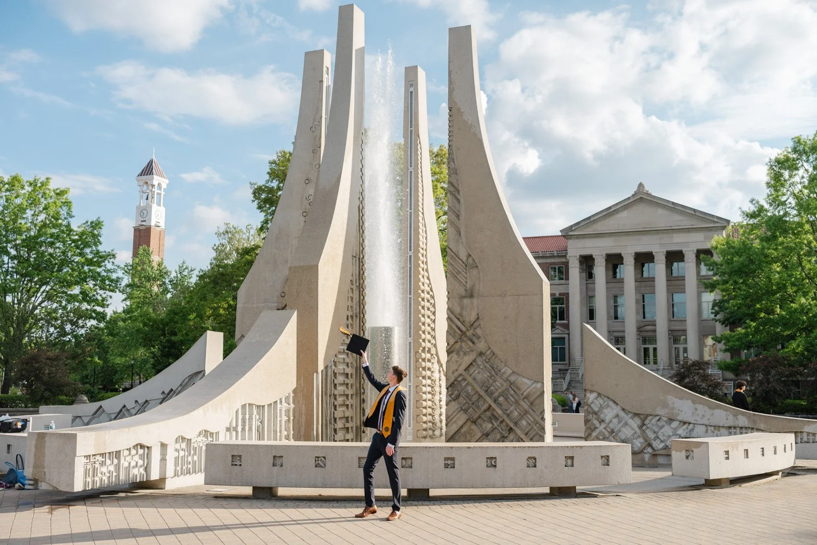 A man raising his Purdue Graduation Cap to cheer for his Purdue Graduation photos.  He is standing in front of the Purdue Engineering fountain with the Purdue Bell Tower and Hovde Hall in the background.  Photo by Heather Corbin Photography.