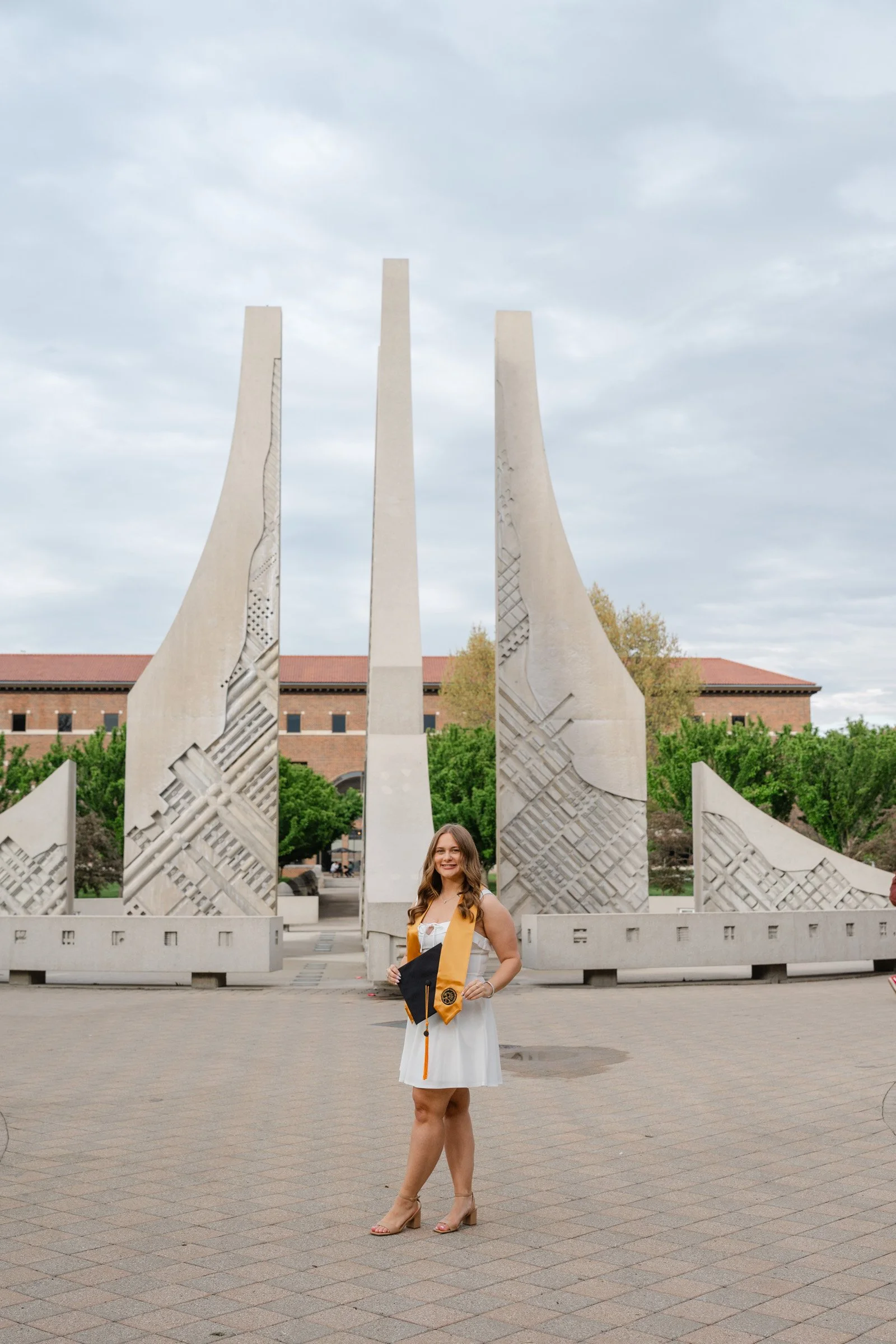 Female Purdue Graduate in white dress with Purdue gold stole and graduation cap standing in front of the purdue engineering fountain