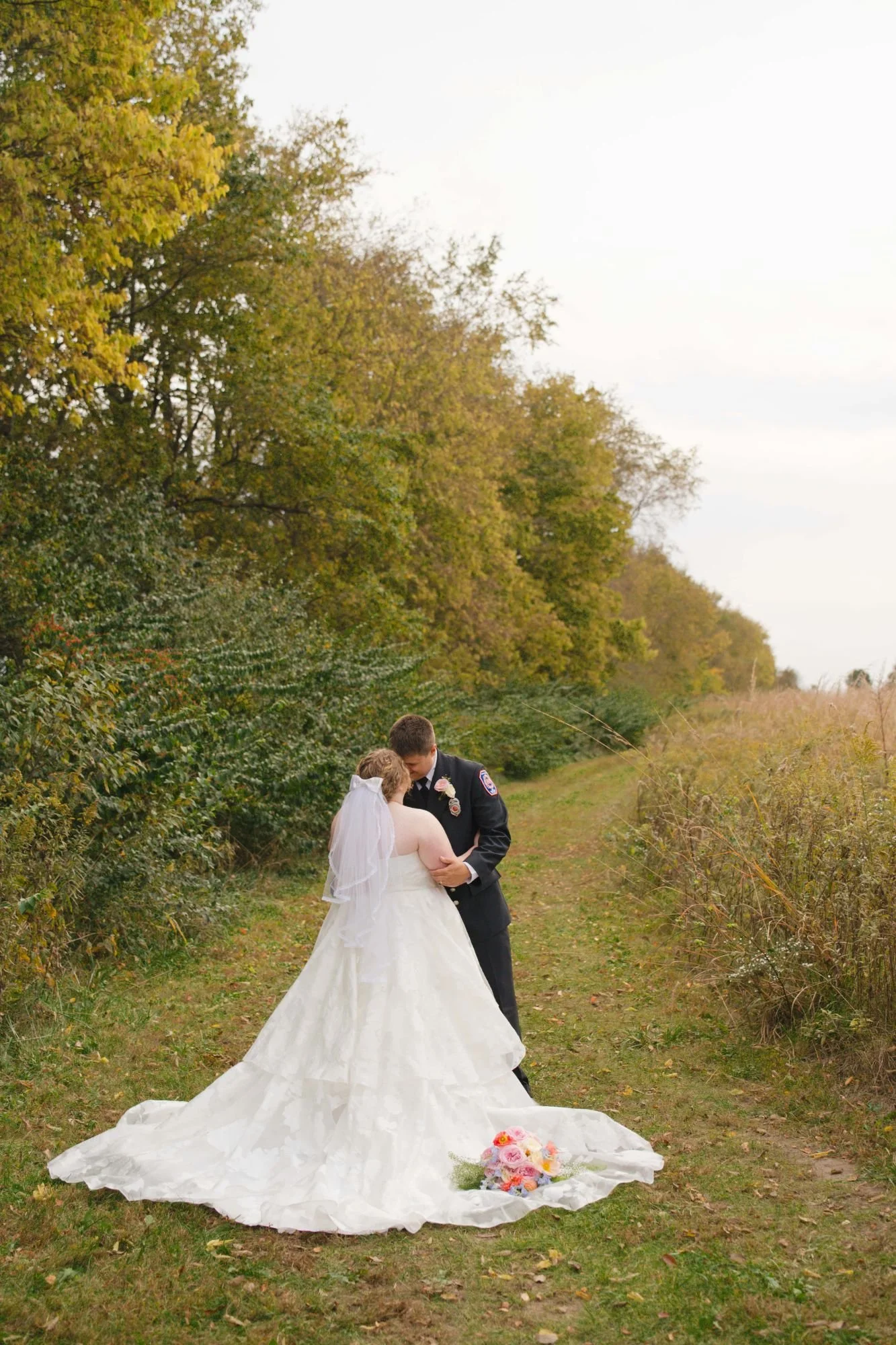 Groom in his firefighter dress uniform facing the camera while the bride is facing him and her full wedding train is spread out behind her.  Grooms is pulling the bride in for a kiss.