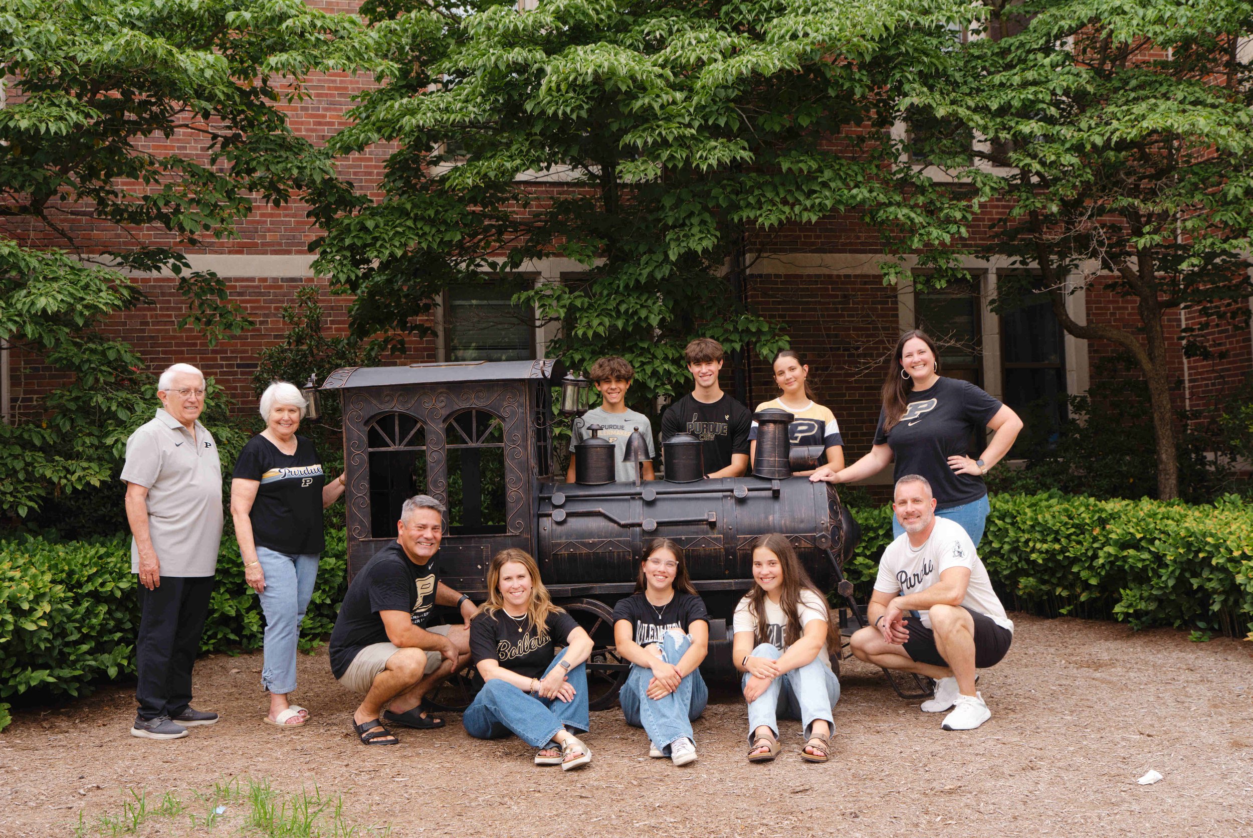3 generations of family posing near a metal train on the Purdue West Lafayette campus during their family photo session.  Everyone is wearing Purdue shirts.