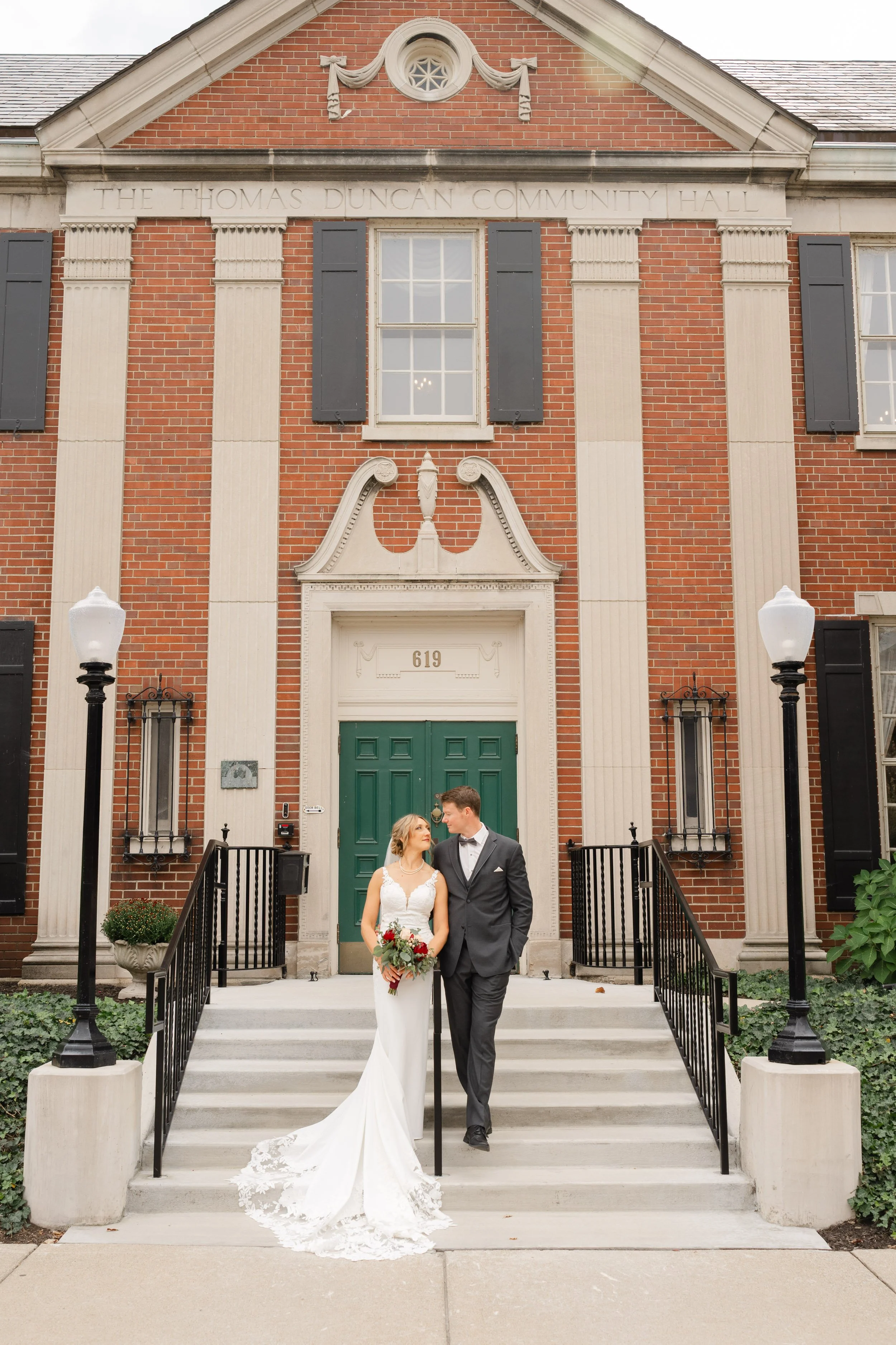 Bride and Groom standing on the stairs in front of Thomas Duncan Hall in Lafayette, Indiana.  Couple is looking at each other.  Bride is holding red and white bridal bouquet.  Photo by Heather Corbin Photography.