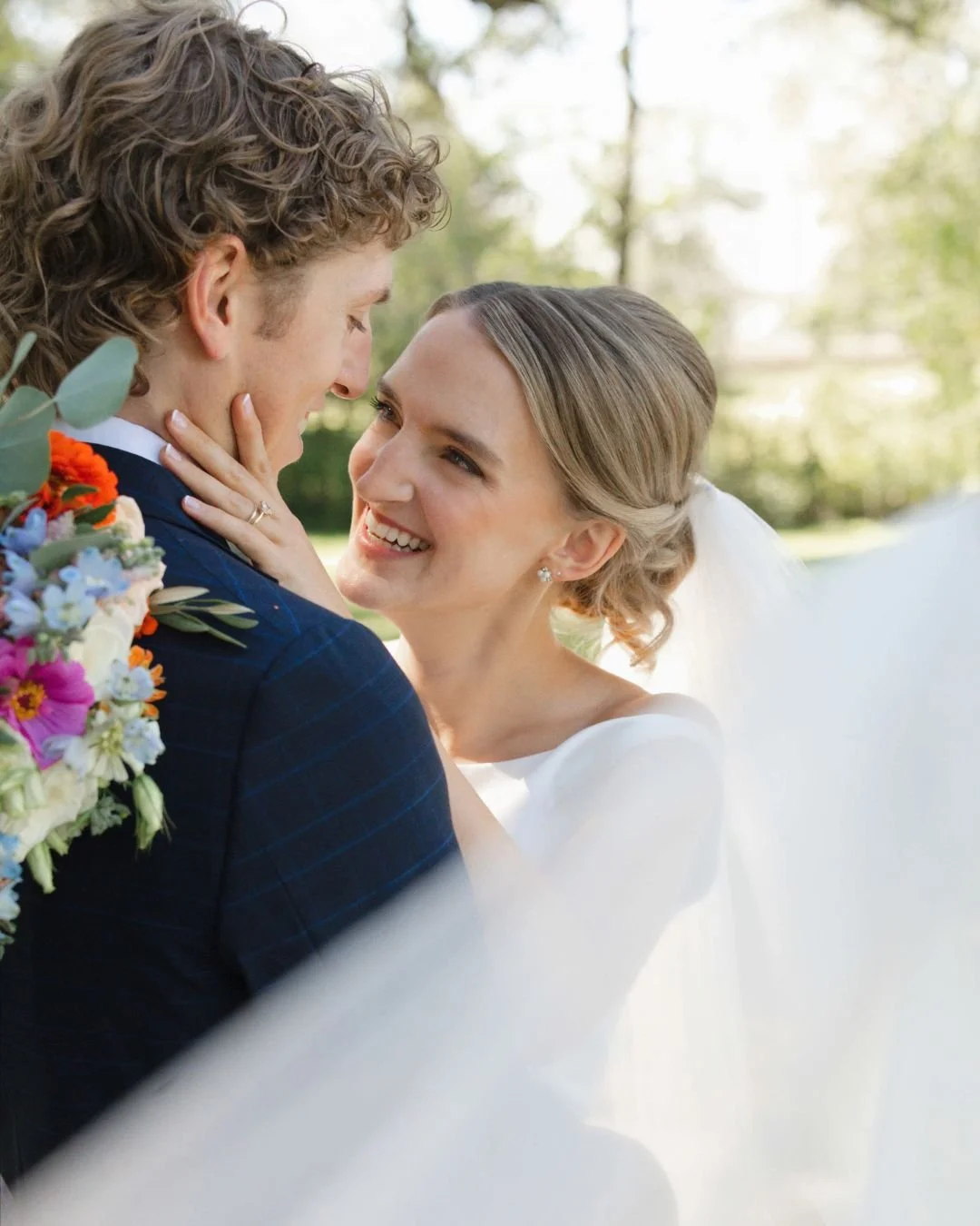 Bride and groom facing each other. Bride's left hand is touching grooms right cheek.  The wedding veil is swooping in front of them.