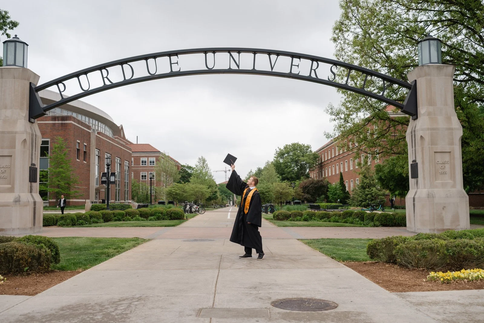 Purdue (male) graduate in a cap and gown standing under the Purdue Arch tossing his cap up in the air.