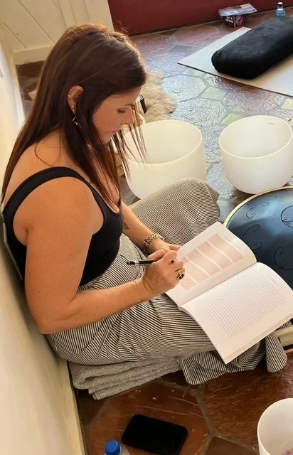 Woman sitting on the floor reading a book with singing bowls and a blanket behind her.