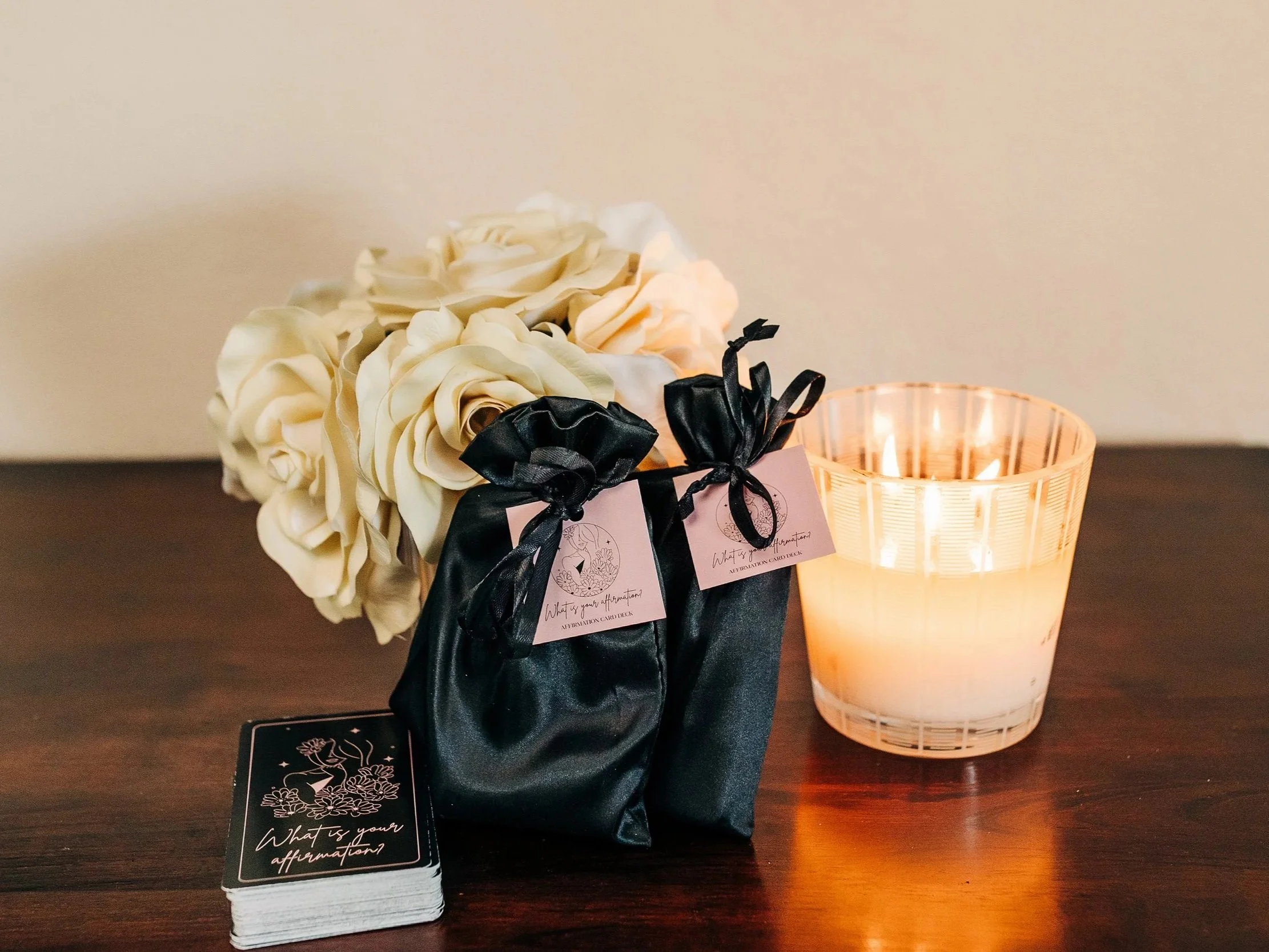 A bouquet of white roses tied with black satin ribbons, a glass candle holder with a lit candle, and a deck of affirmation cards on a dark wooden surface.