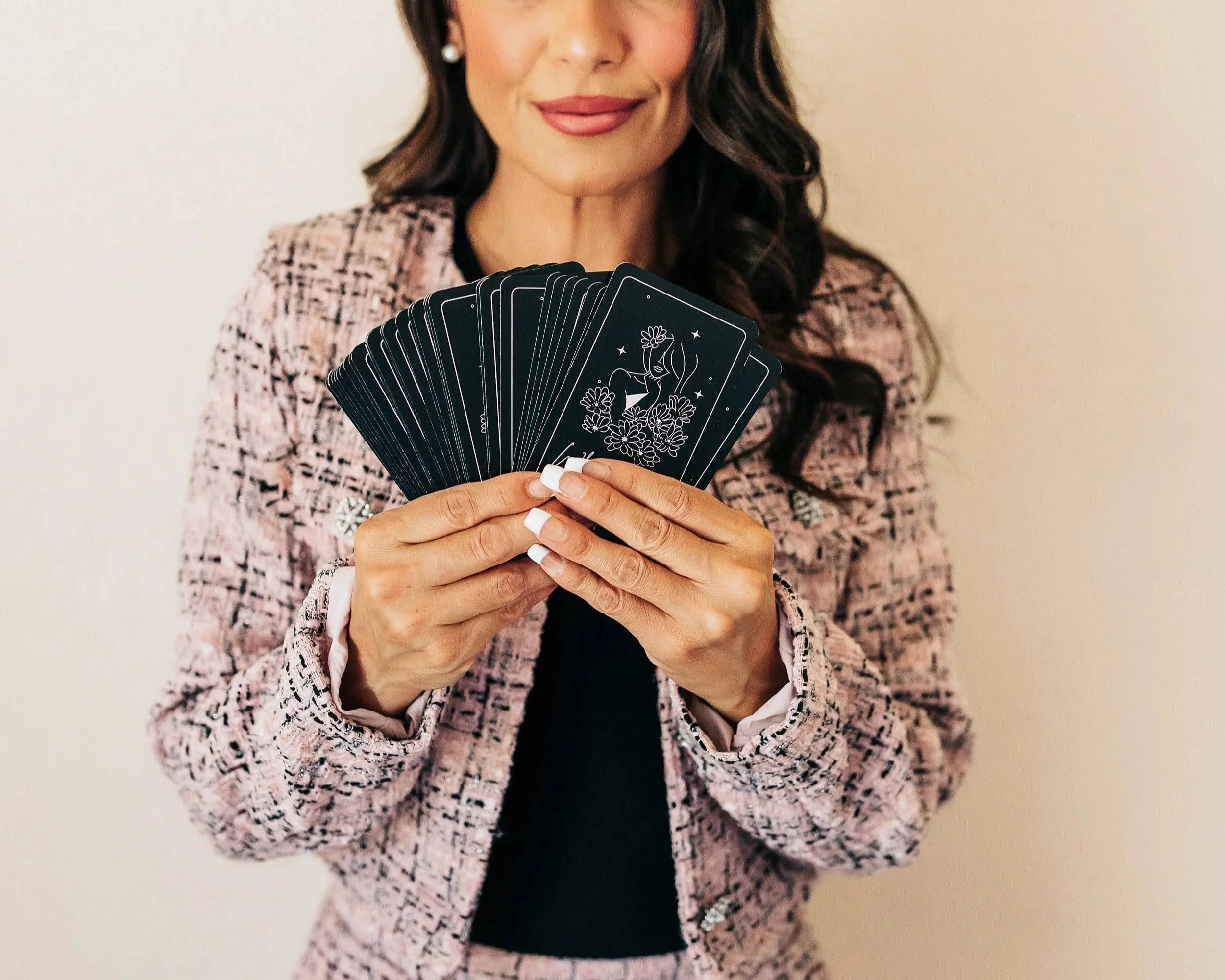 Woman holding a fan of black tarot cards with white line art design in front of her face, wearing a pink patterned blazer.