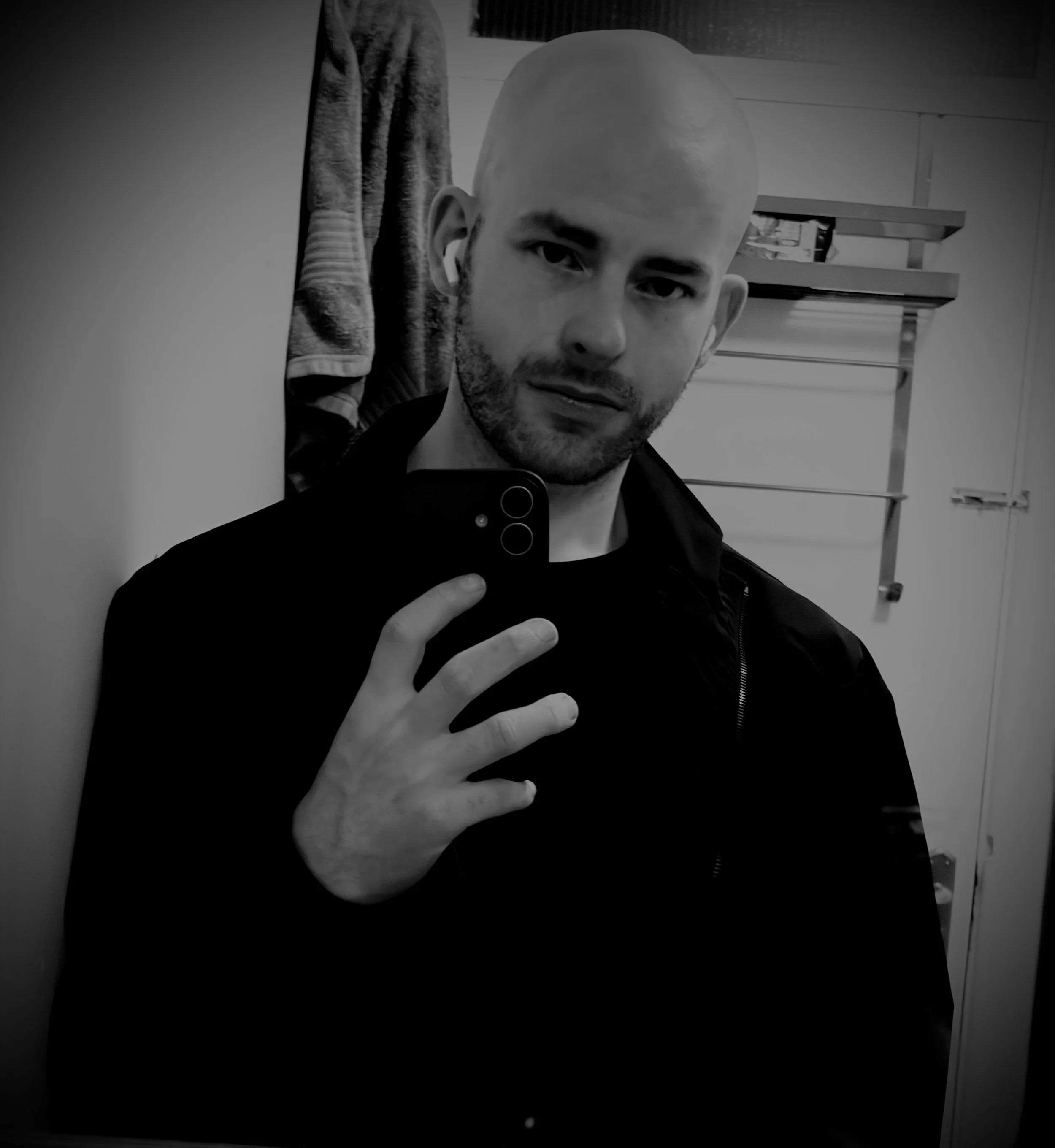 A man taking a mirror selfie in black and white, showing part of his face, hand, and a smartphone. He has a bald head, beard, and earbuds, and is wearing a dark jacket. The background includes a towel hanging on a hook and metal shelves with various items.