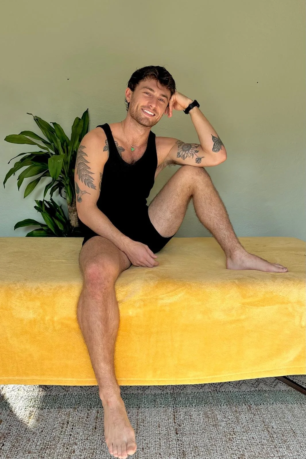 A man with tattoos, black tank top, and black shorts sitting on a yellow cushioned surface with one leg extended and the other bent, posing with his head resting on his hand, smiling, with a green houseplant and plain wall in the background.