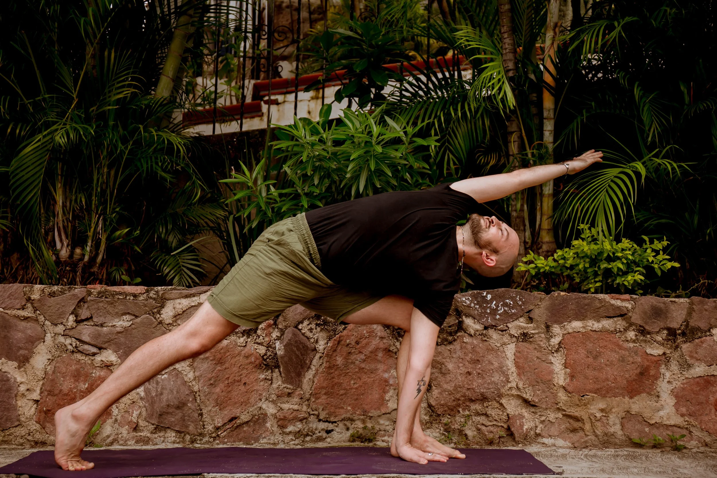 A man performing a yoga pose outdoors on a yoga mat, surrounded by lush green plants and trees.
