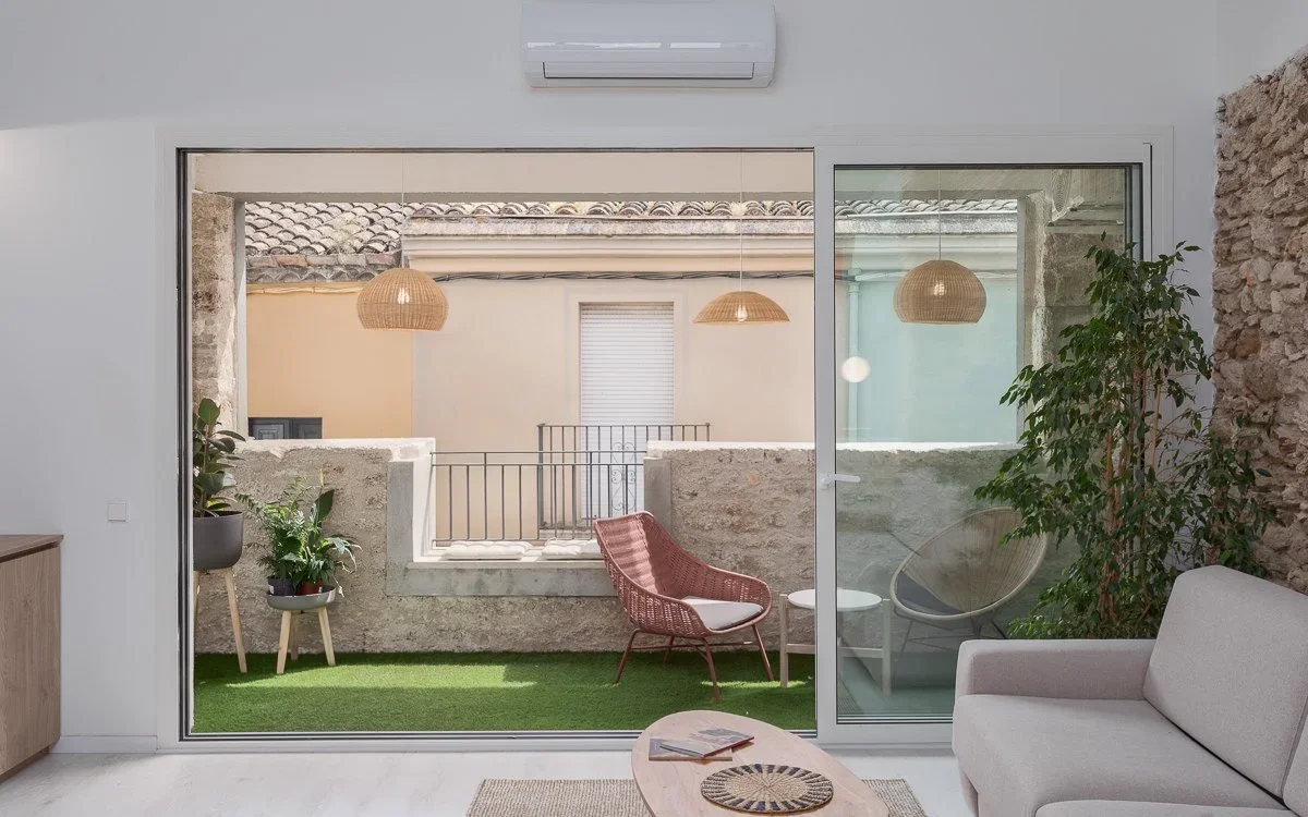 Innenhof eines renovierten Ferienhauses in Banyoles, Girona, mit freigelegten Steinwänden, modern-minimalistischem Design, Terrasse mit Kunstrasen und Korbmöbeln.