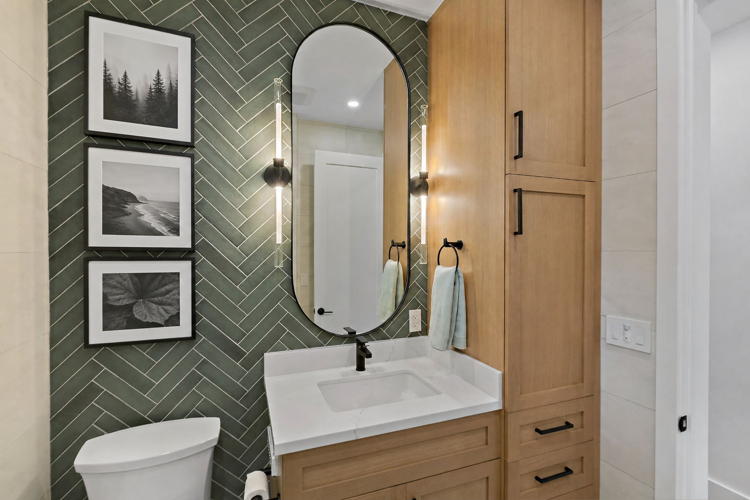 Modern bathroom with a black-framed oval mirror over a white vessel sink, black faucet, wooden cabinetry, black towel rings with towels, green tile wall in a herringbone pattern, black and white artwork of nature scenes, and beige tiled walls.