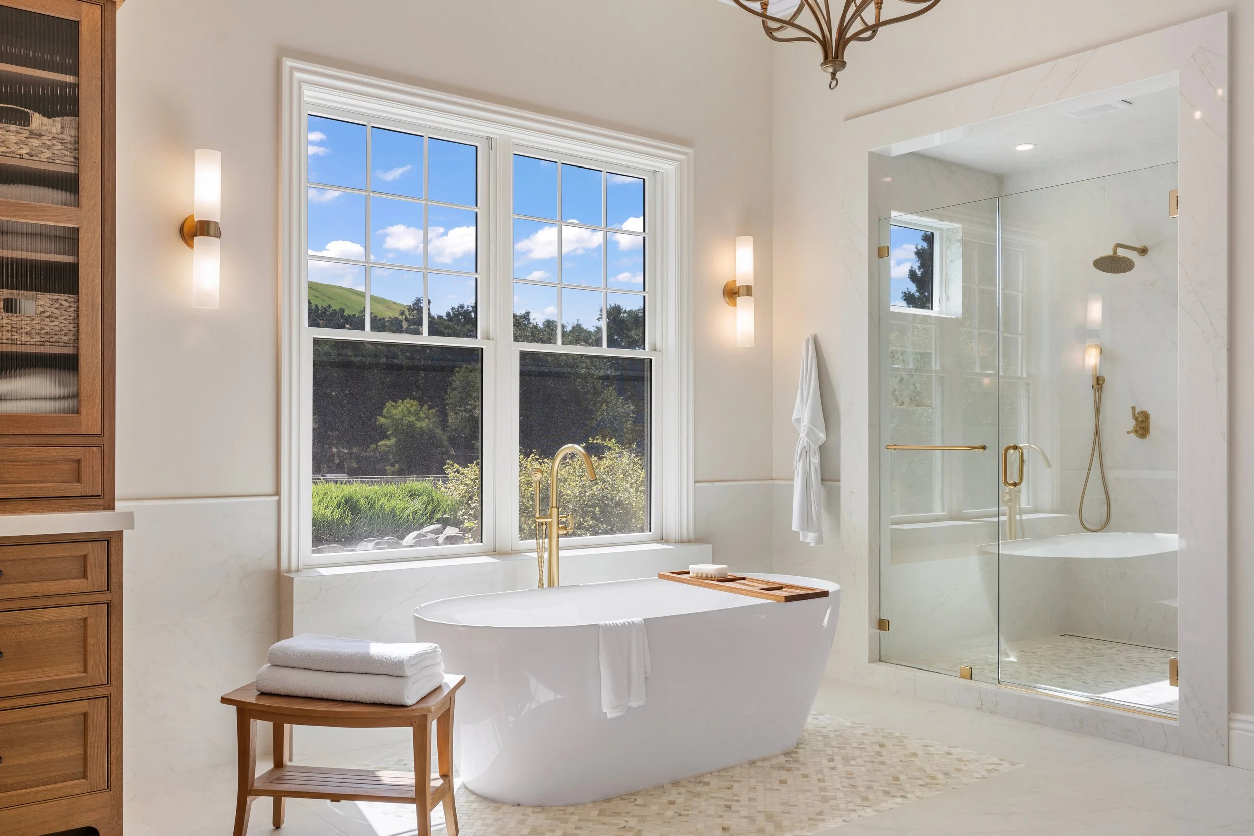 Photo of a modern bathroom featuring a freestanding bathtub with a wooden tray holding a brush and a towel, a glass-enclosed shower area with gold fixtures, a large window showing green hills and blue sky with clouds, and warm wall-mounted lights.