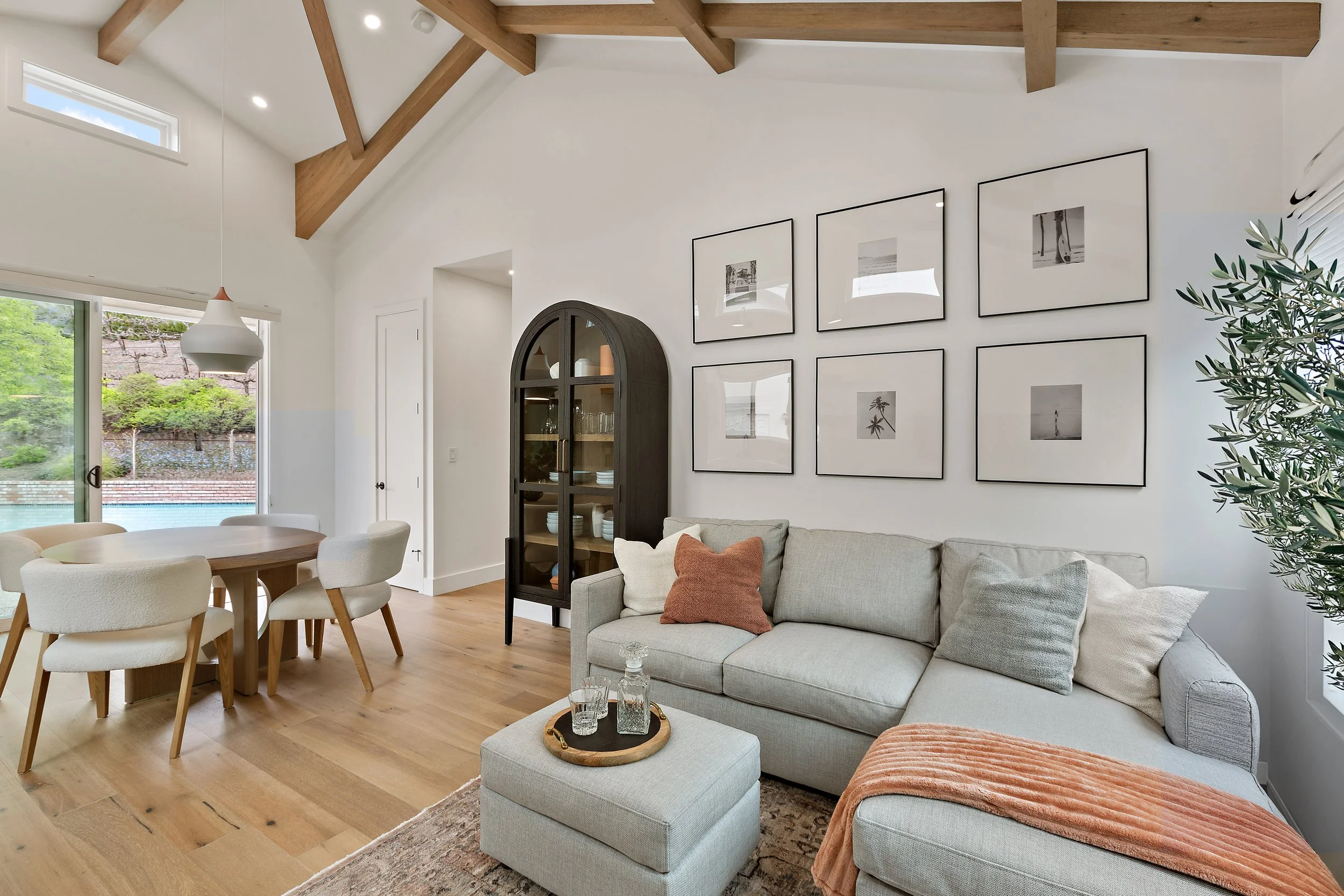 Modern living room with light-colored sofa, framed black-and-white photographs on the wall, wooden ceiling beams, large windows with a view of greenery, dining table with chairs, and decorative items like a cabinet and tray with glasses.