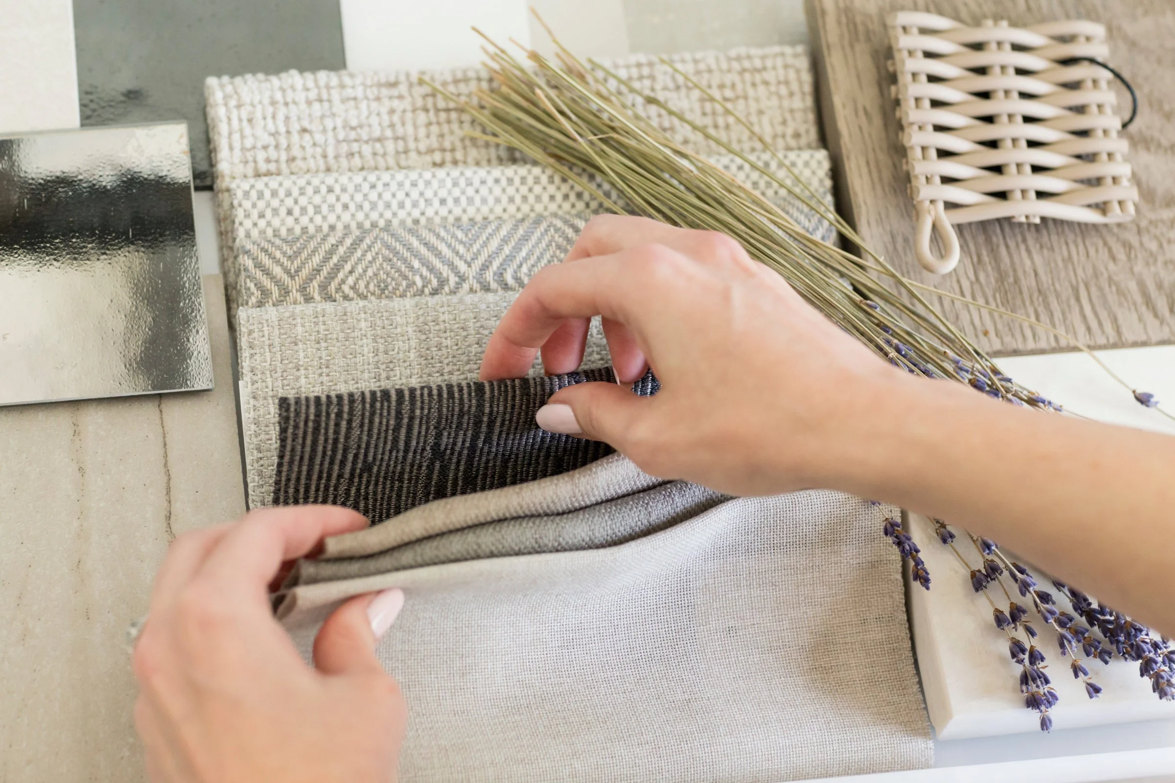Person's hand folding a fabric in beige, grey, and black shades, with a lavender branch nearby on a textured surface.