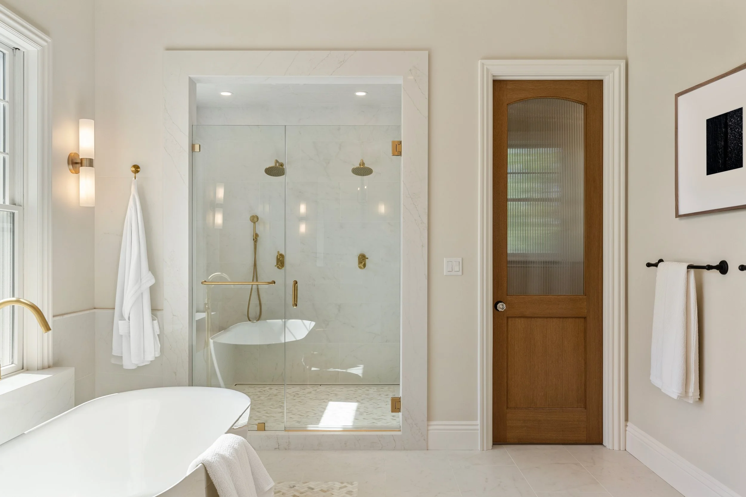 Modern bathroom with a glass-enclosed shower featuring gold fixtures, a white bathtub, a window with sunlight, and a wooden door with textured glass.